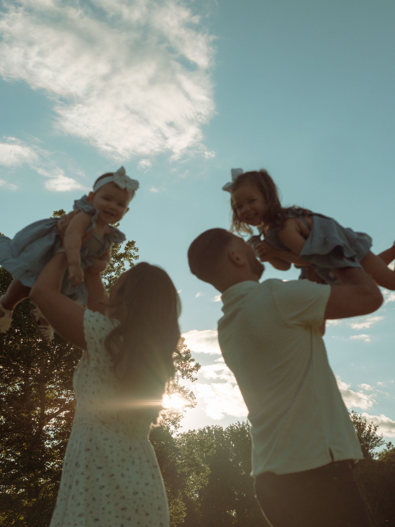 Parents lifting two young daughters in the air against a bright blue sky.