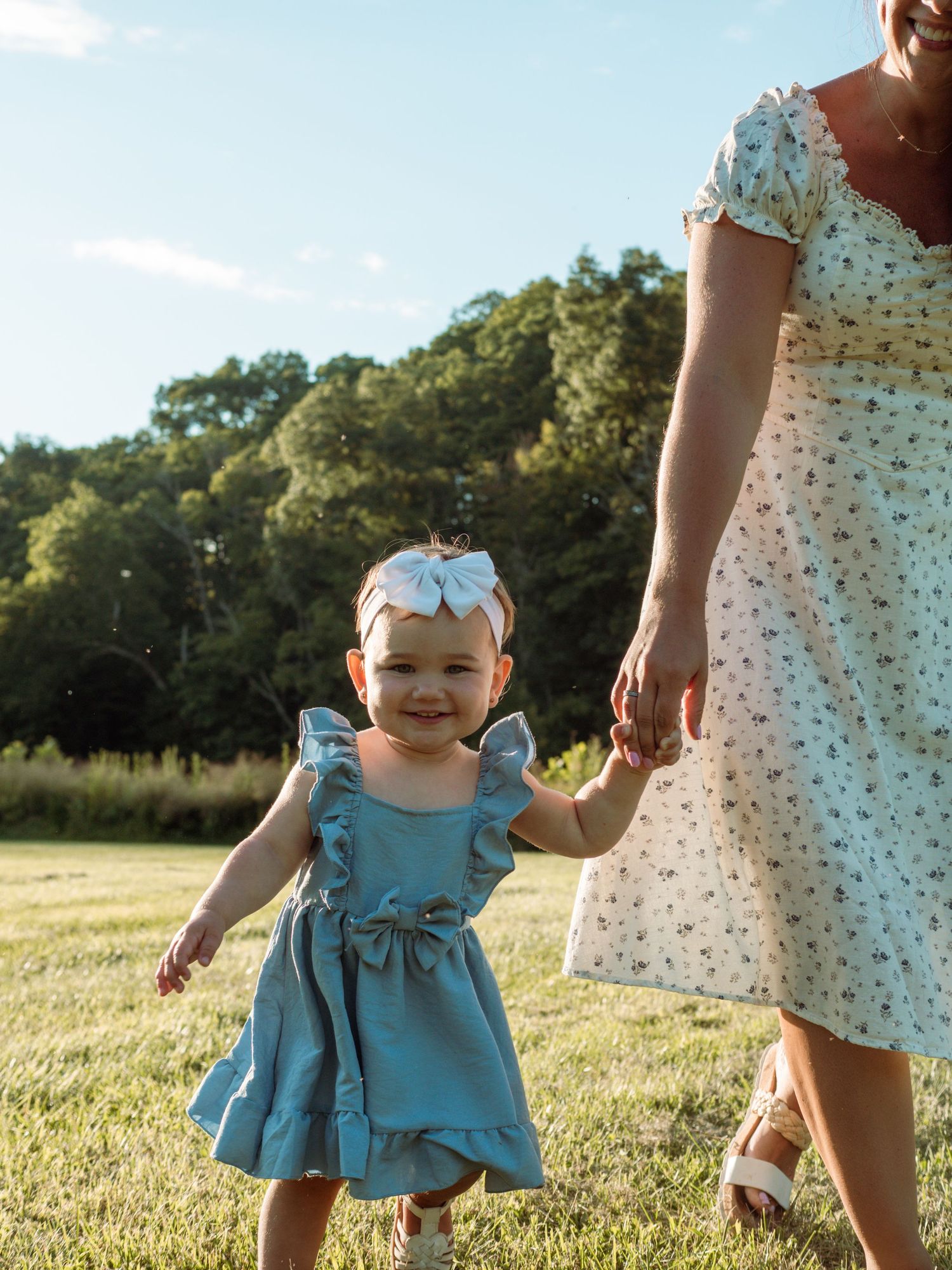 Smiling toddler girl in blue dress holding mother's hand in sunny field.