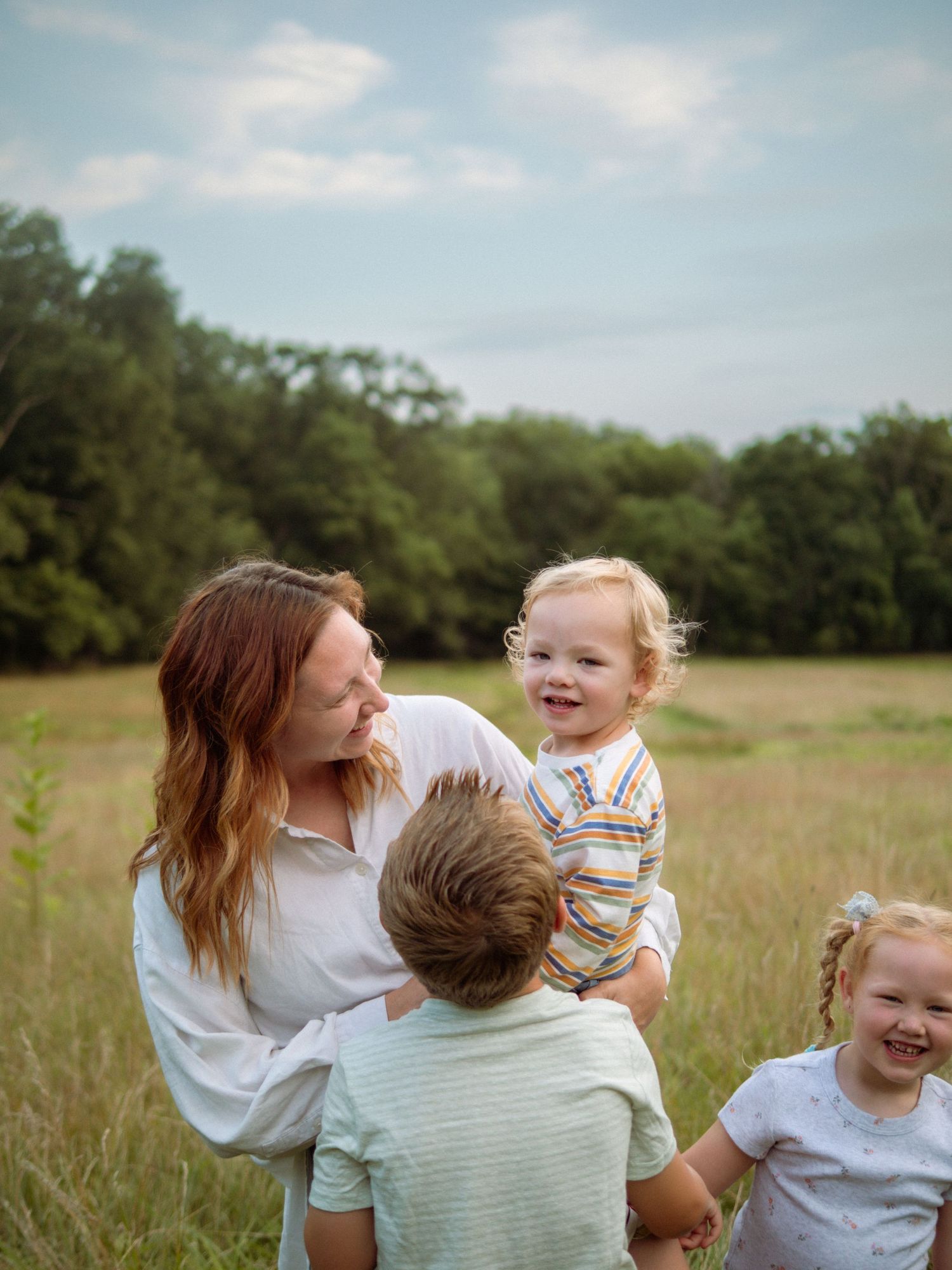 Joyful mother holds laughing toddler in sunny field while two other children stand nearby.