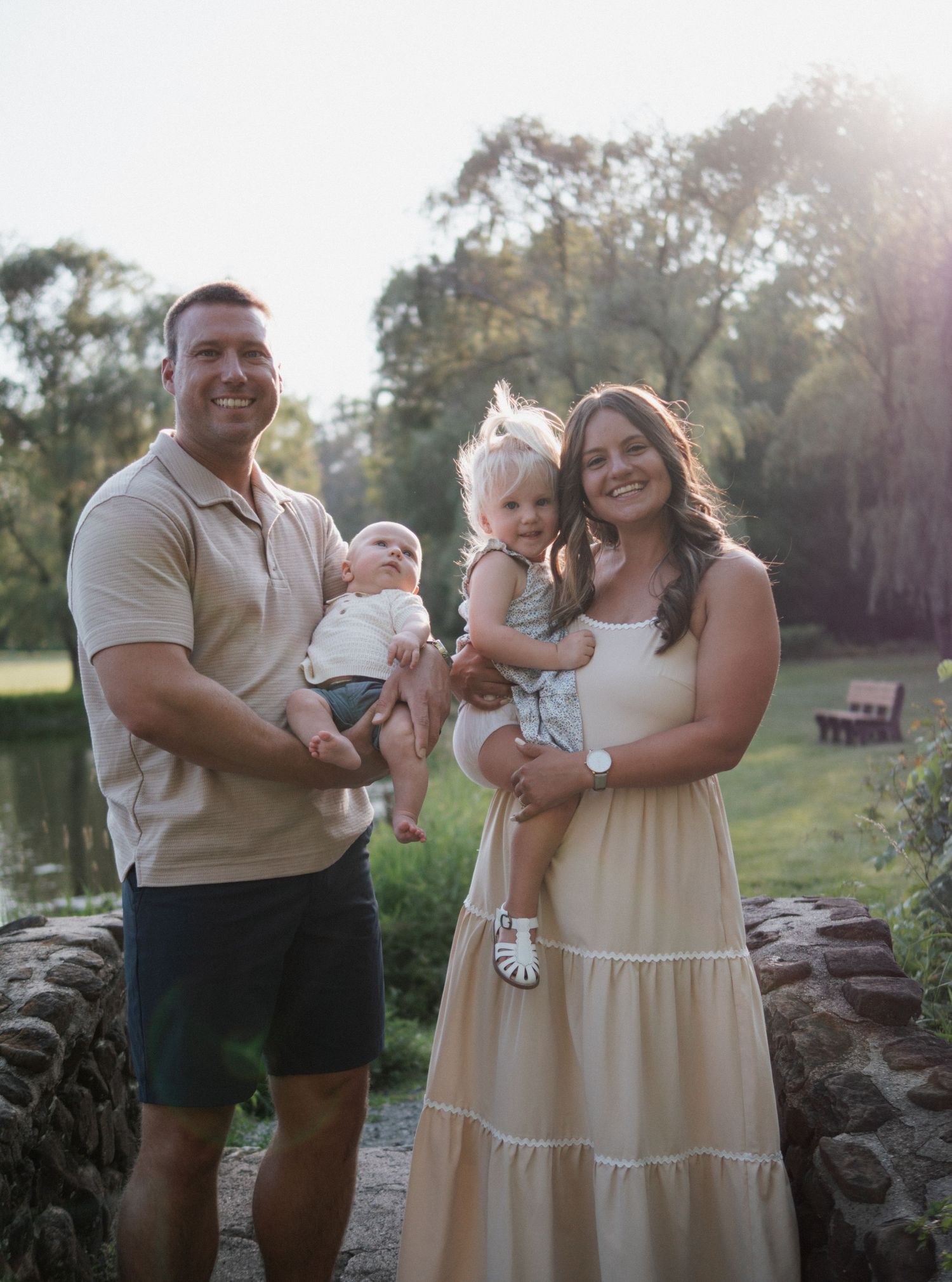 Smiling family of four with infant and toddler posing outdoors in warm sunset light.