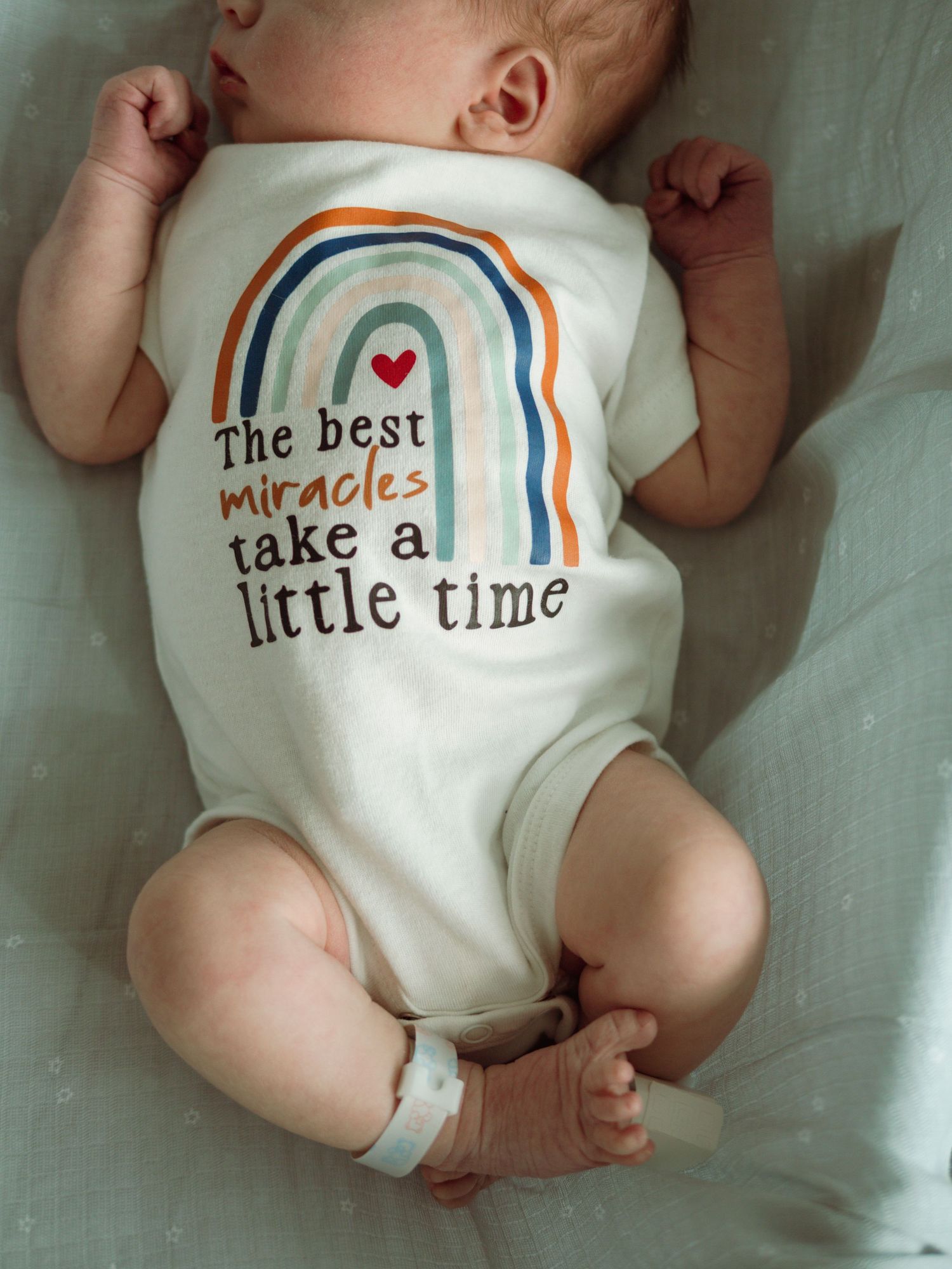 Newborn baby wears onesie reading the best miracles take a little time during hospital photo session.