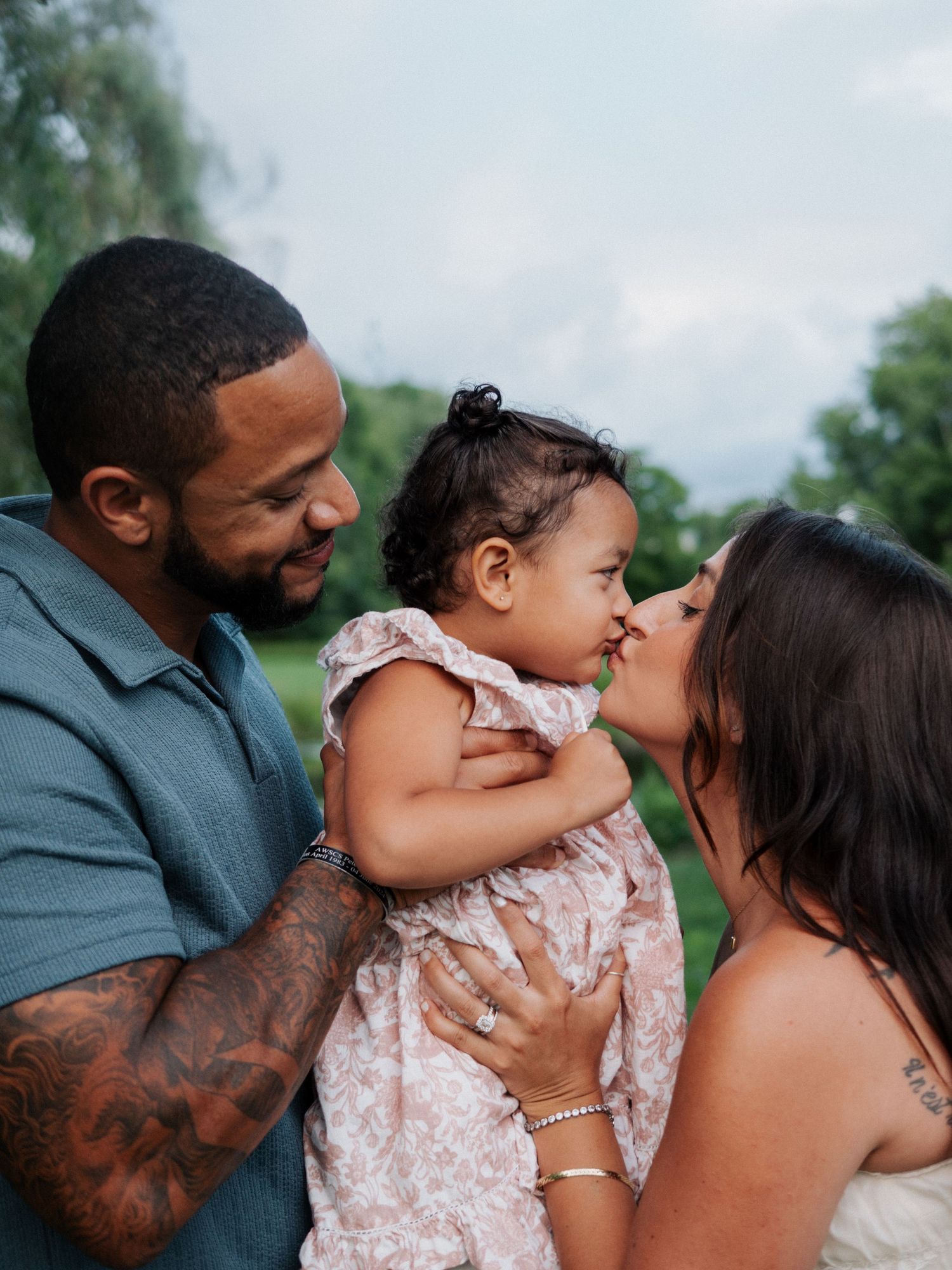 Parents sharing a kiss with their smiling toddler daughter between them outdoors.