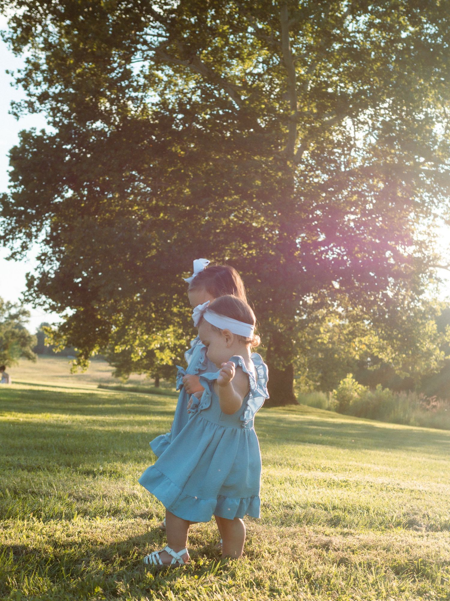 Two young sisters in matching blue dresses playing in a sunlit park.