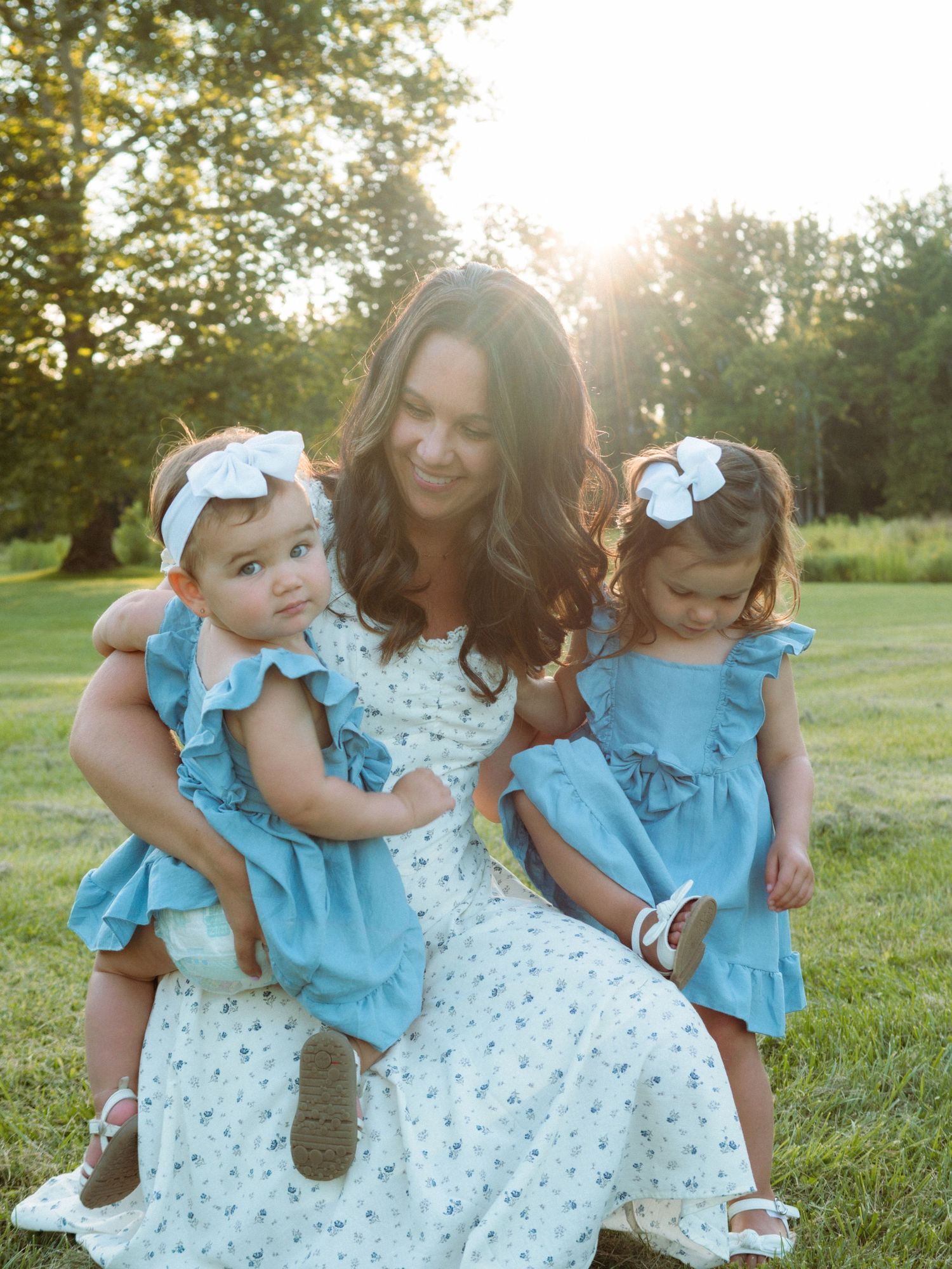 Mother sitting outdoors with two toddler daughters in matching blue ruffle dresses in golden sunlight.