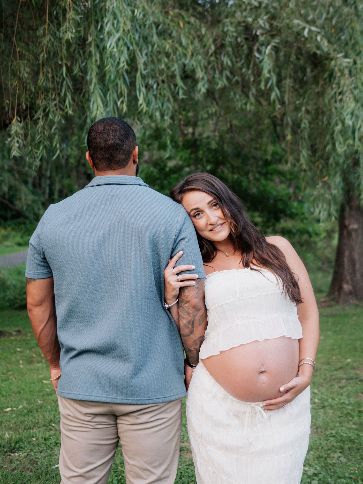 Pregnant woman in white dress leans on partner outdoors under willow tree during maternity photo session.