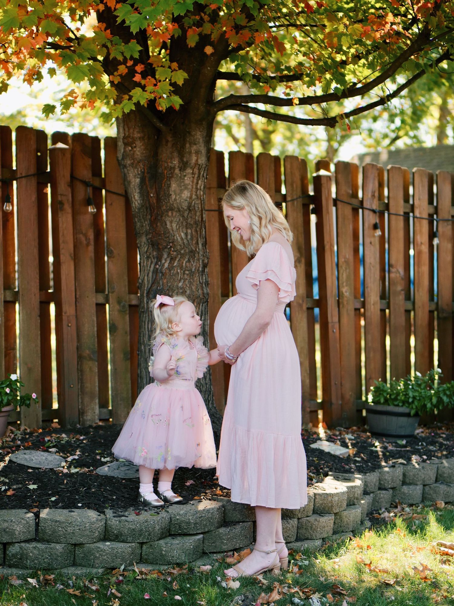 Pregnant woman in pink dress standing with young daughter in matching pink outfit under autumn tree.