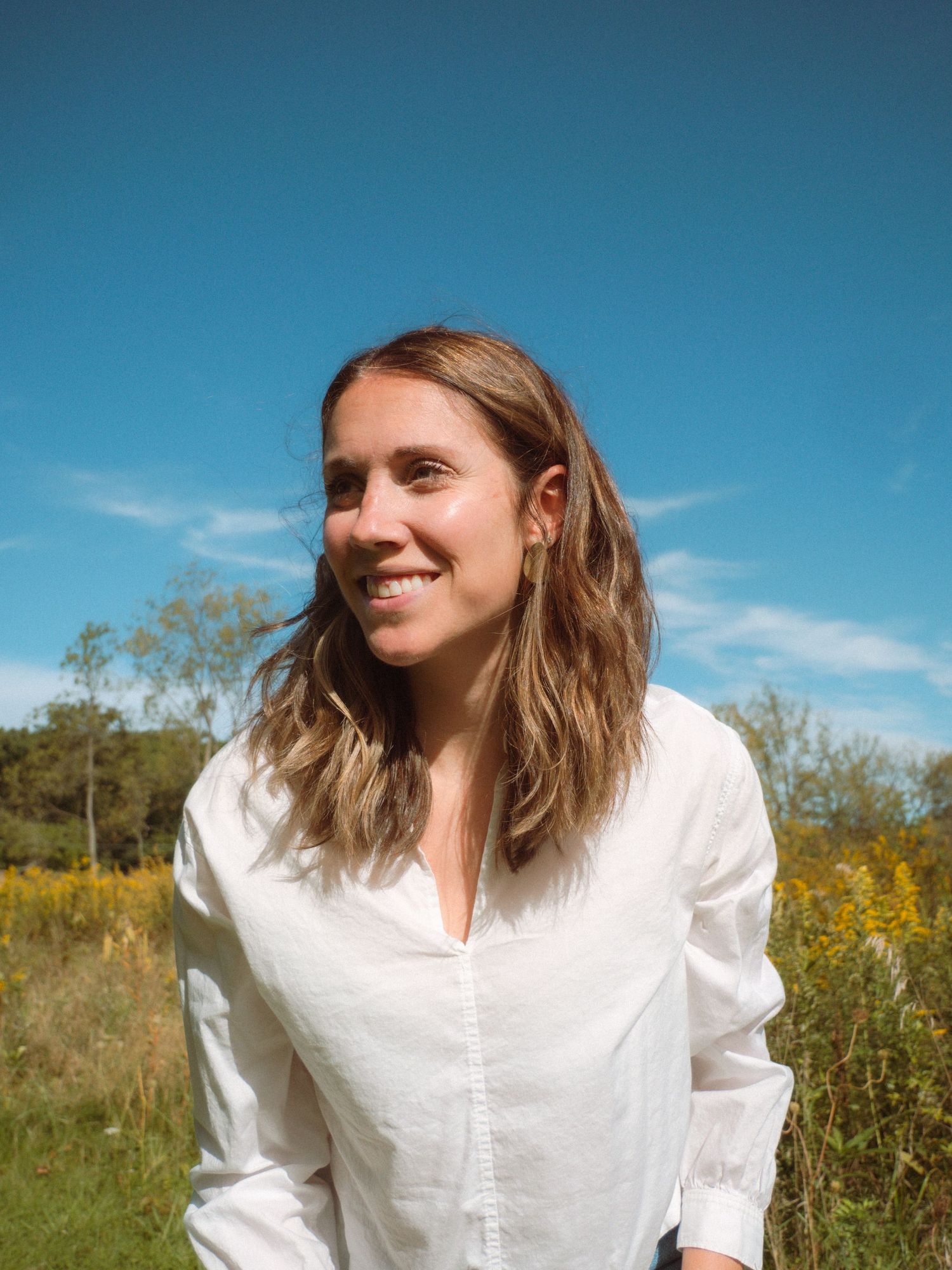 A happy woman with shoulder-length hair smiles outdoors in a bright sunny meadow.