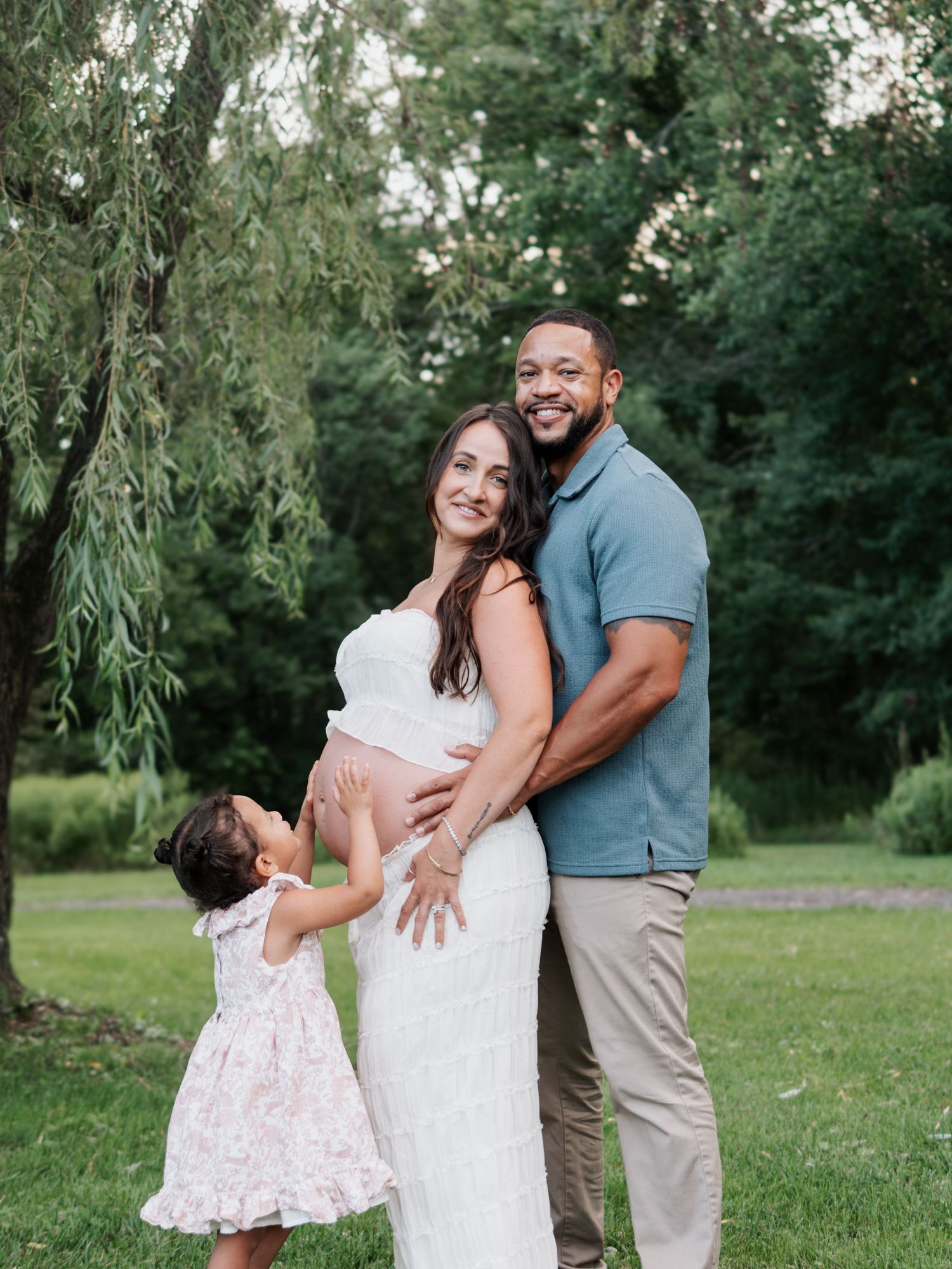 Pregnant woman in white dress with husband and toddler daughter posing for maternity photos in lush green park.