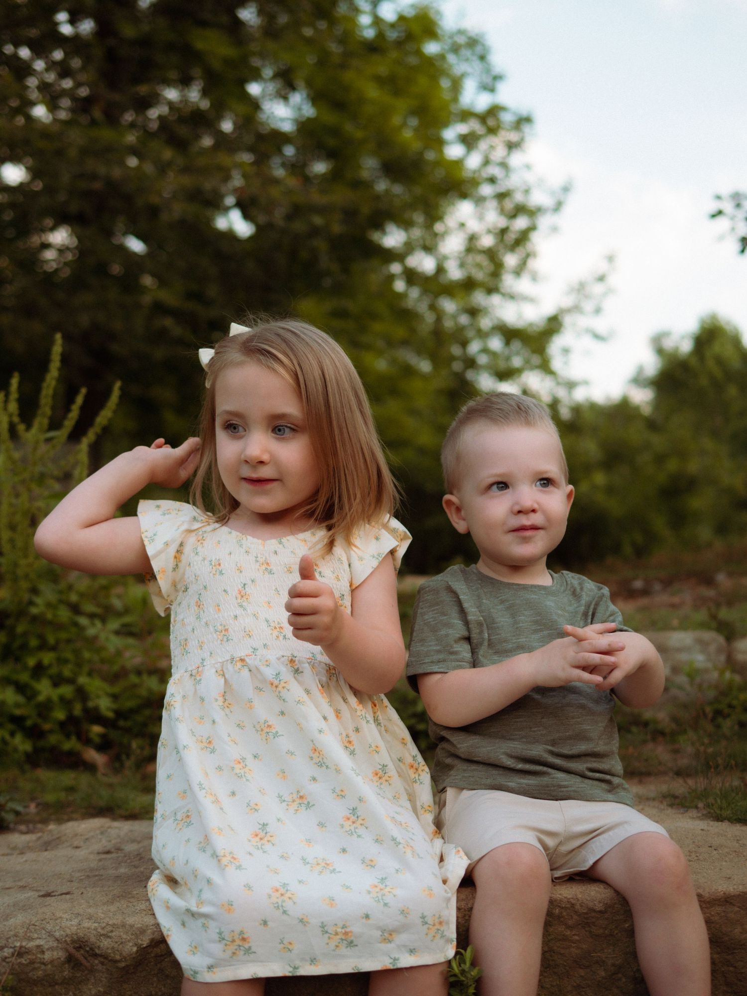 Young girl in floral dress and toddler boy in green shirt sit together outdoors.