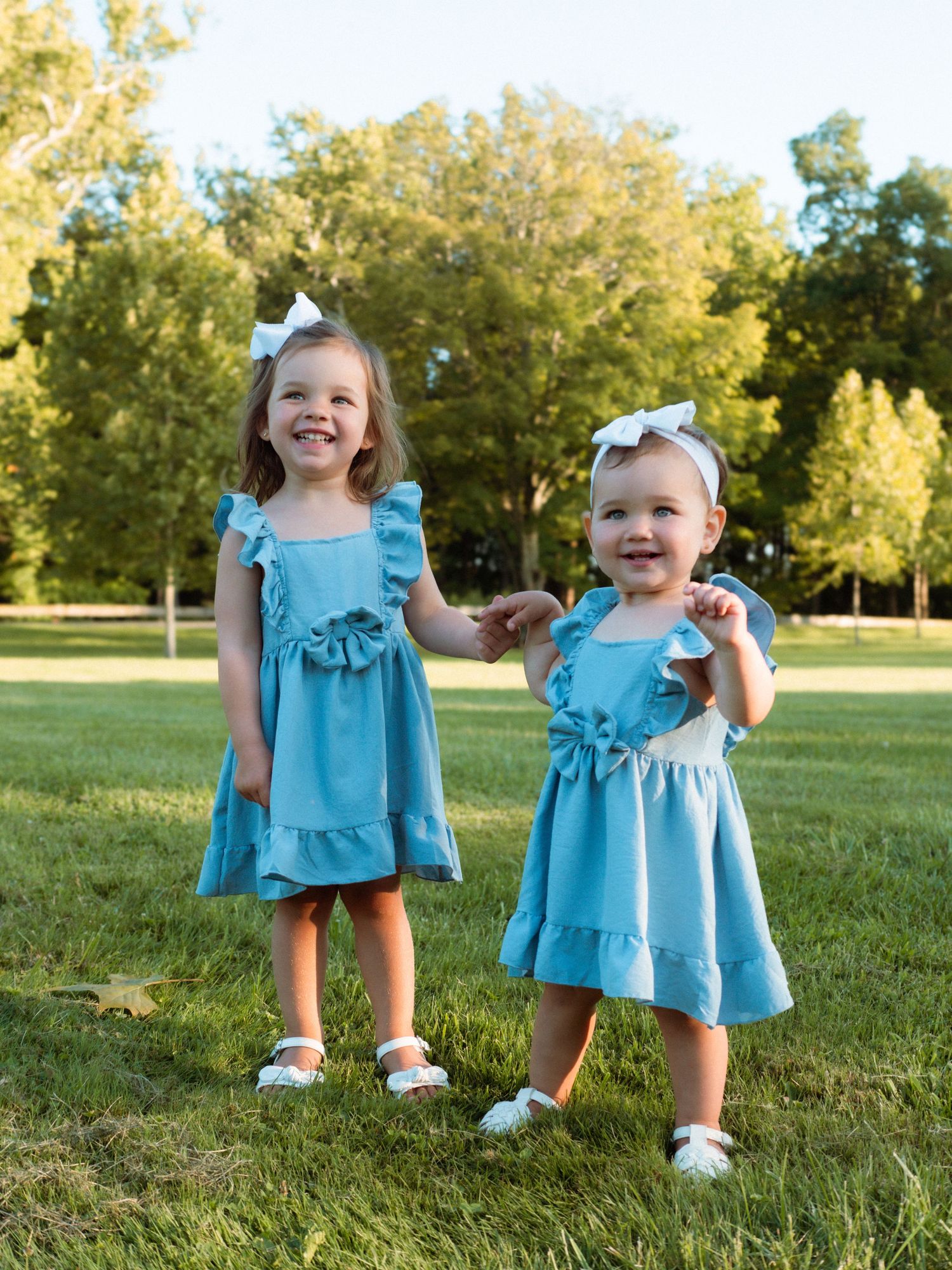 Two little sisters in matching blue dresses smile and hold hands in a sunny park.