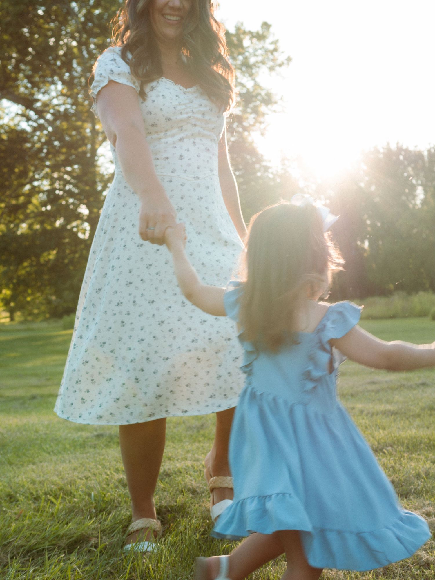 Mother and daughter in matching dresses dancing in a sunlit green field.
