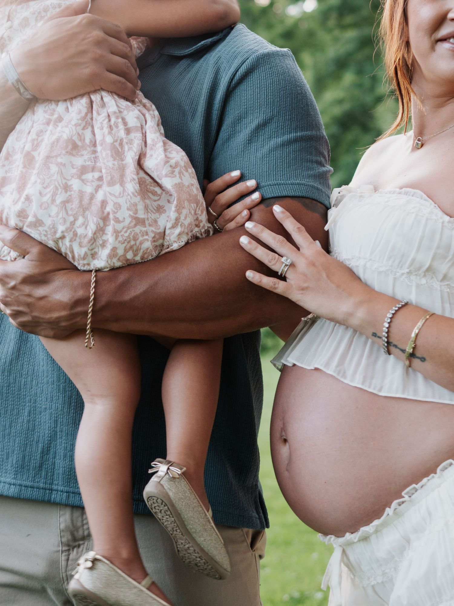Close-up of expectant parents holding toddler daughter during outdoor maternity photo session in garden.