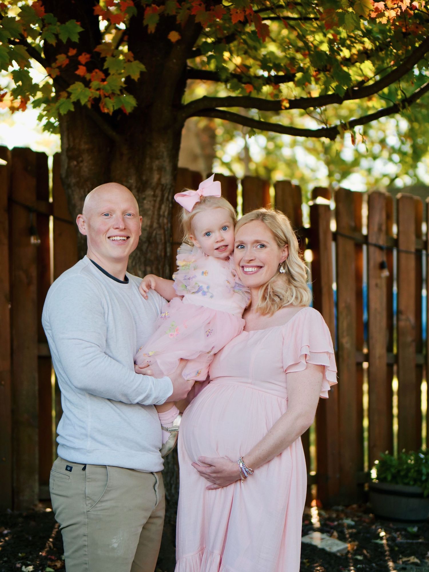 Expecting family posing outdoors with toddler daughter under a colorful autumn tree.
