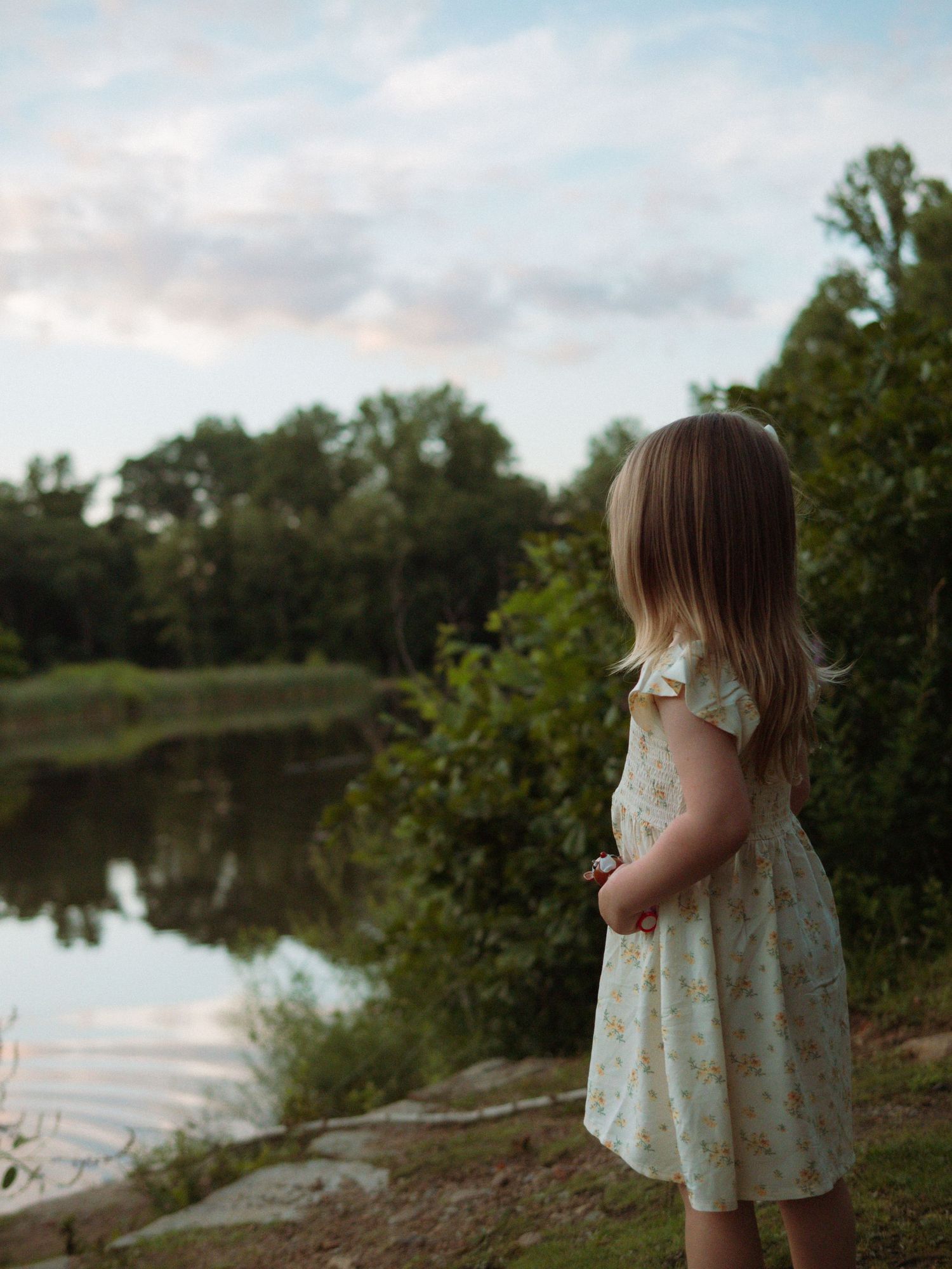 Young girl in floral dress gazes at a peaceful pond surrounded by lush green trees.