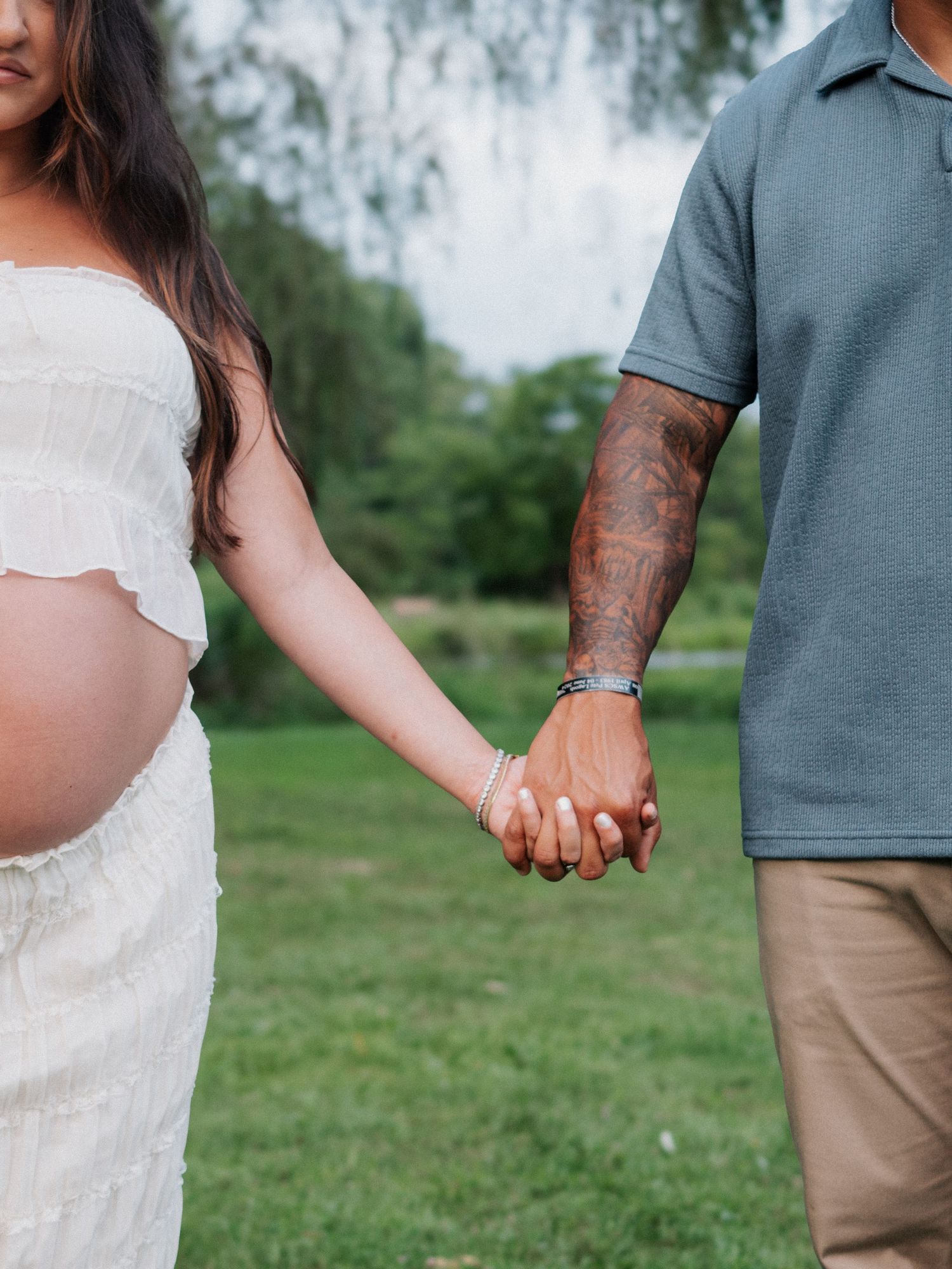 Close-up of pregnant woman and tattooed partner holding hands during outdoor maternity photo session.