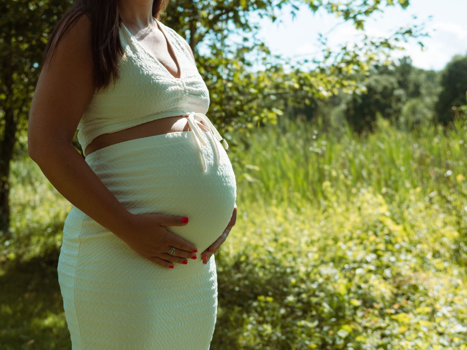 Pregnant woman in mint green dress cradling baby bump surrounded by lush green foliage.