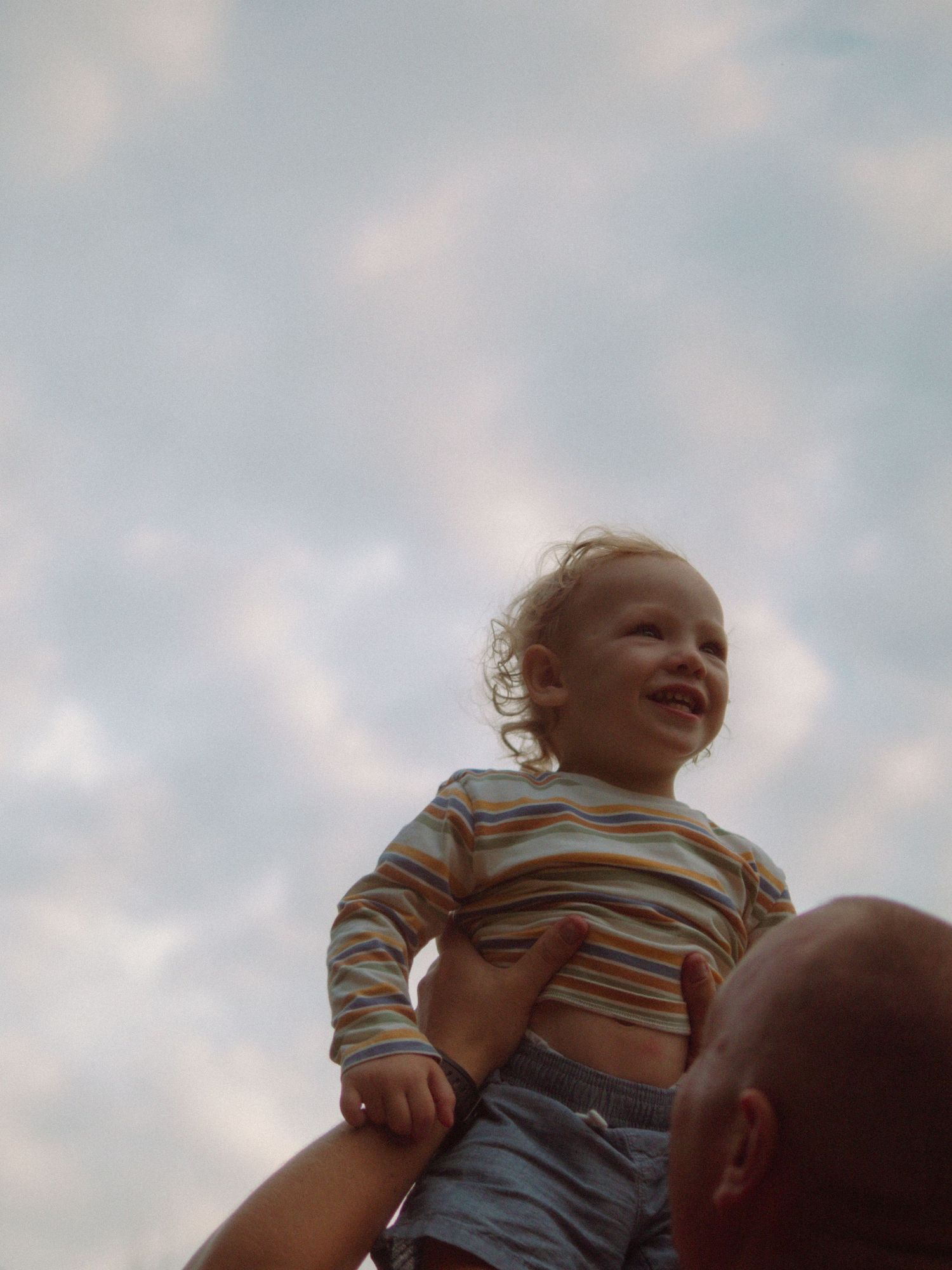 Happy toddler lifted high against a cloudy sky, wearing a colorful striped shirt.