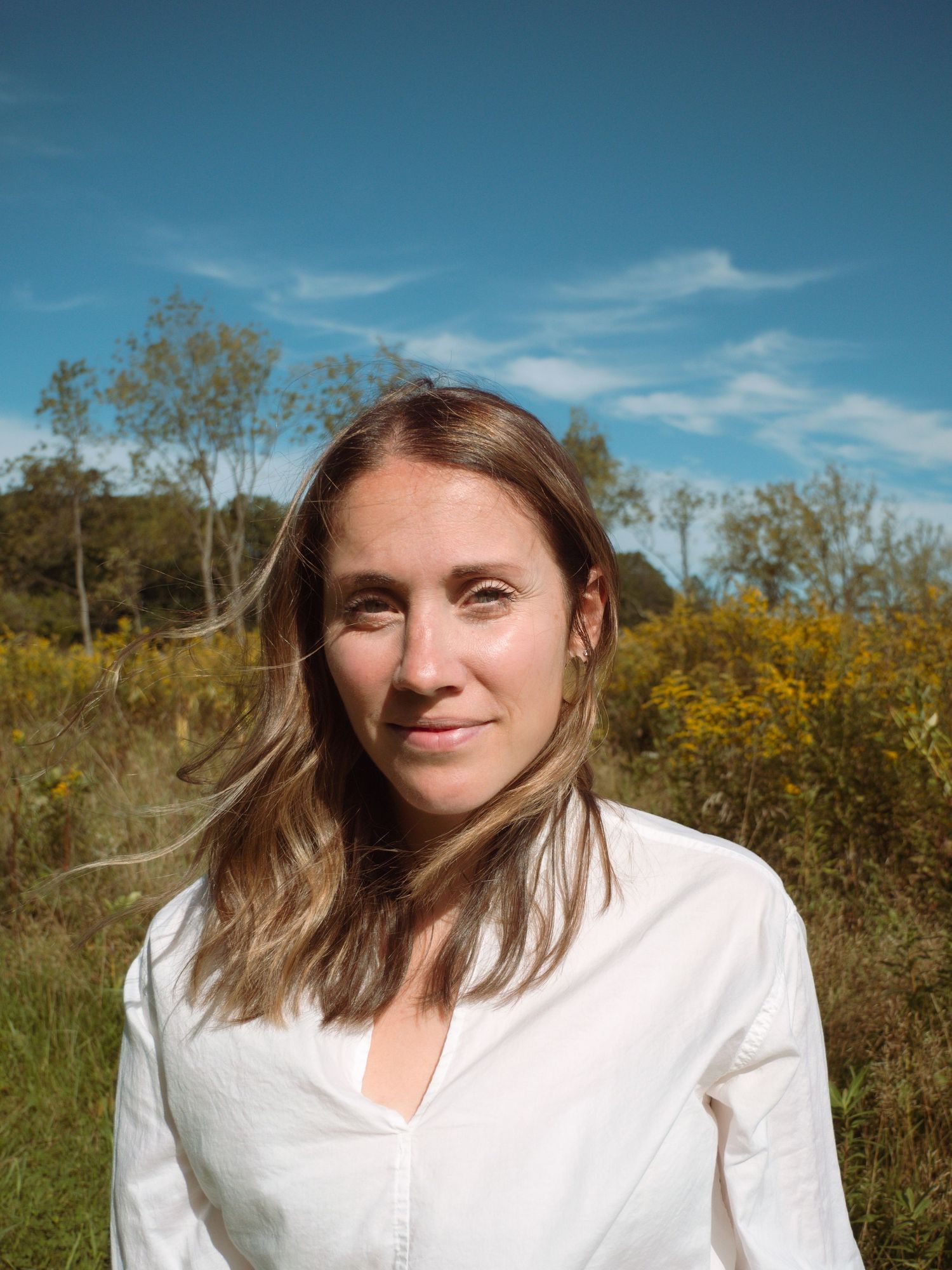 Smiling woman with light brown hair wearing white shirt standing in sunny field with blue sky background.