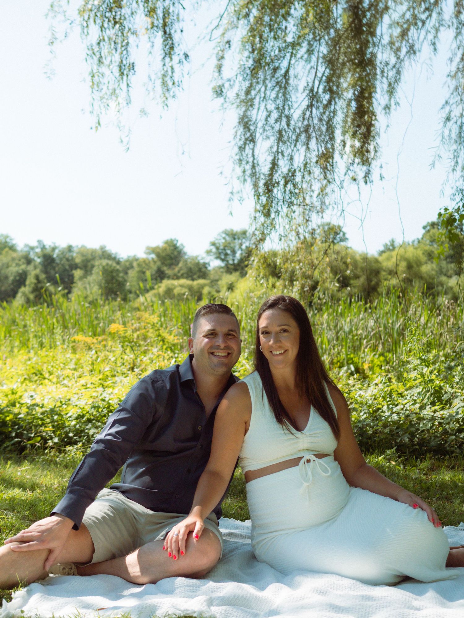 Pregnant couple sitting together on blanket under weeping willow tree on sunny summer day.