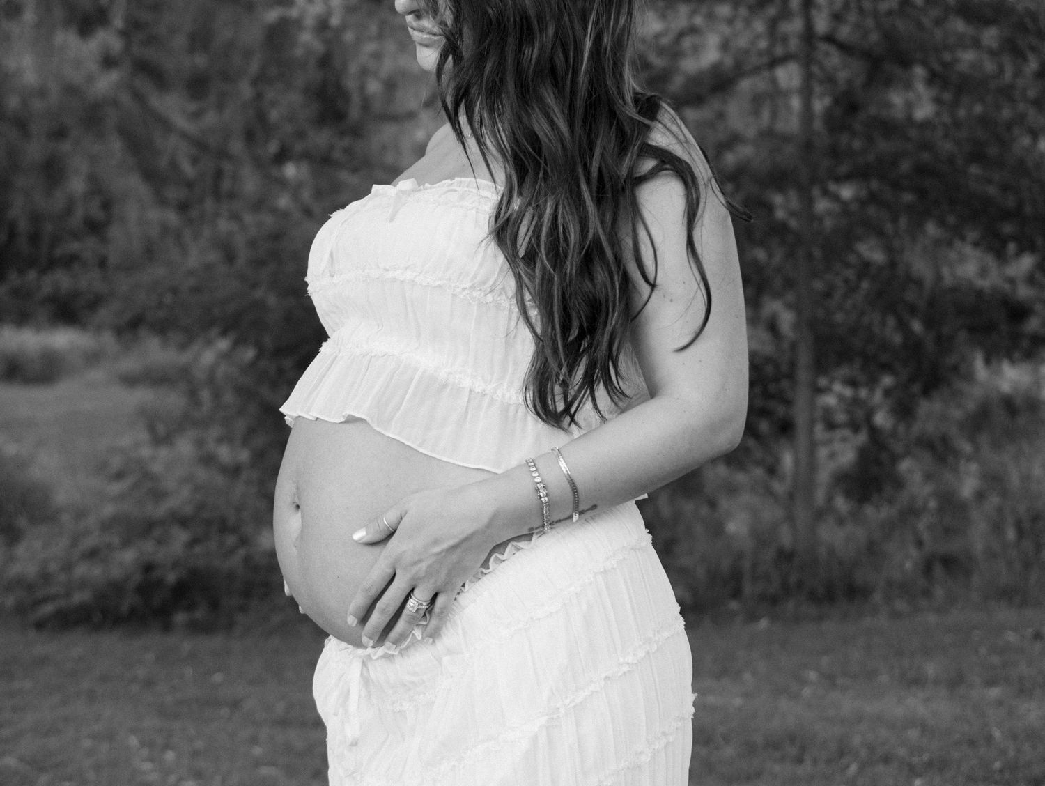 Black and white maternity portrait of pregnant woman in white dress holding baby bump in garden.