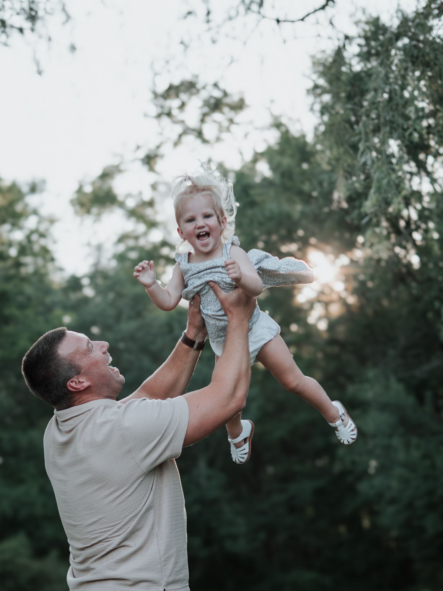 Father joyfully lifting laughing toddler girl into the air in a sunlit forest.