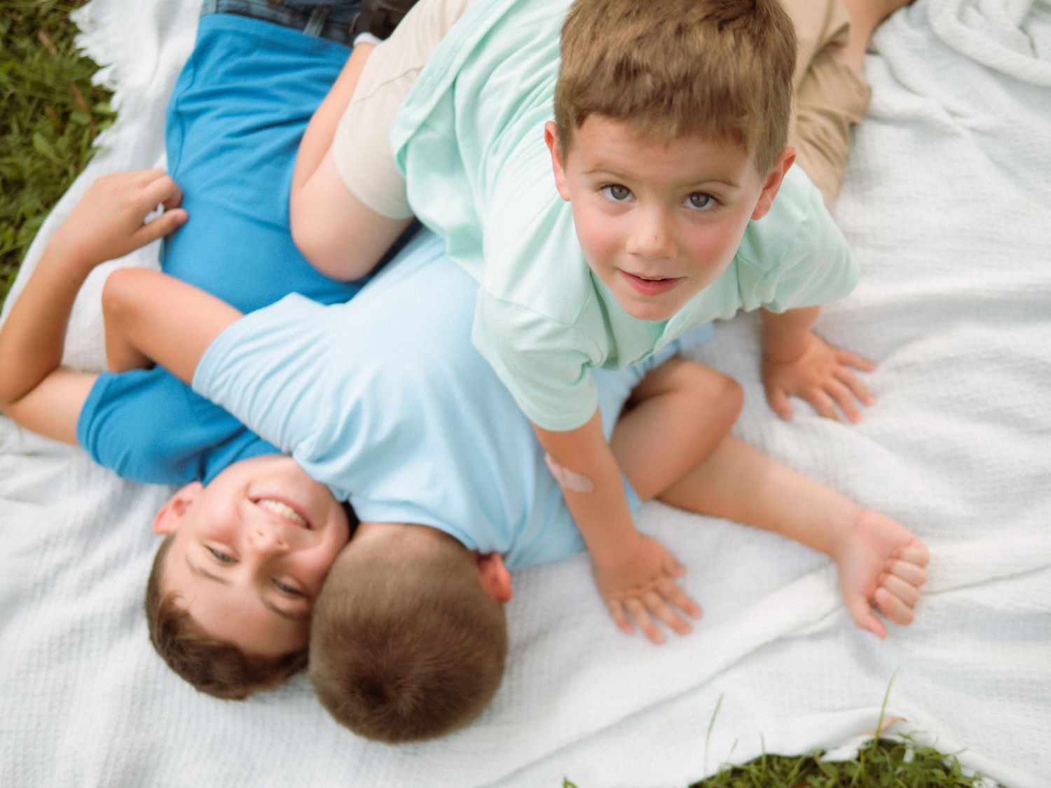 Three boys in light blue shirts laugh and play together on a white blanket outdoors.