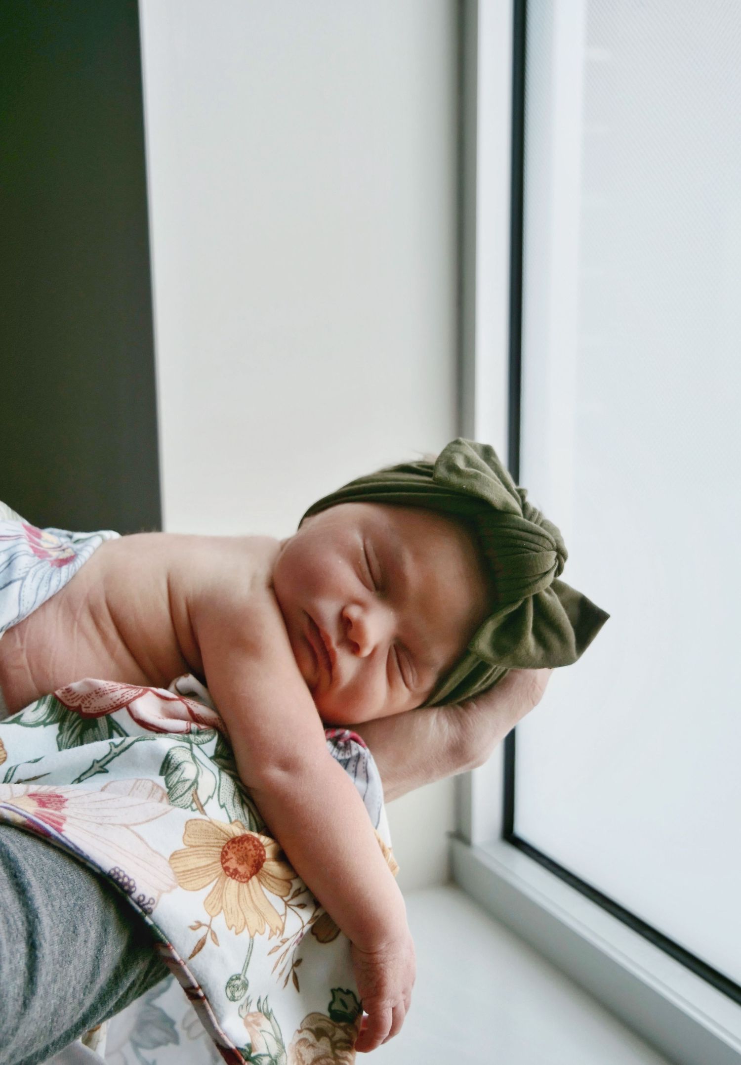 Sleeping newborn baby wearing an olive green bow headband, cradled in an adult's arms.