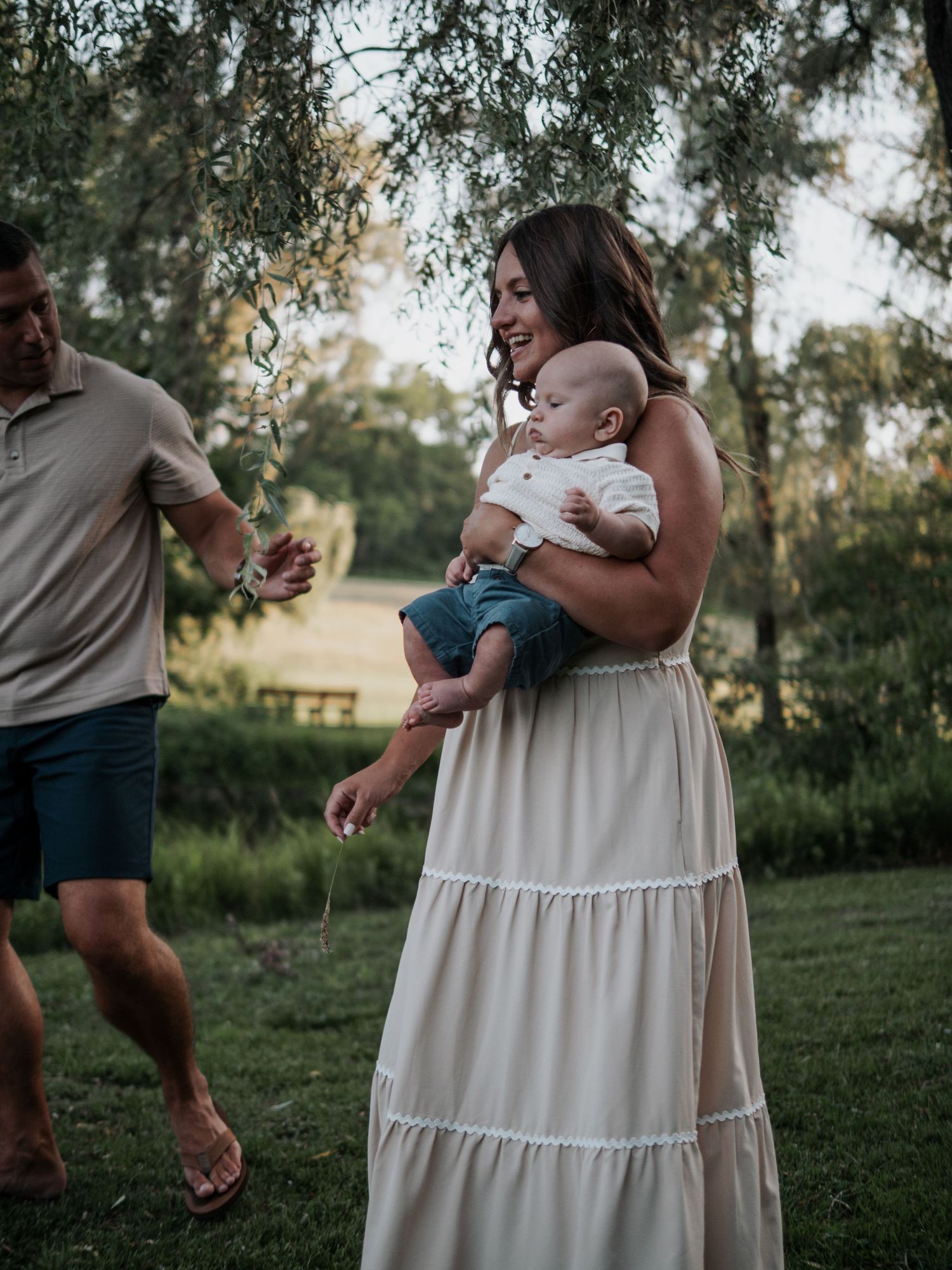 Smiling mother in white maxi dress holds baby boy outdoors while father walks nearby in lush green park.