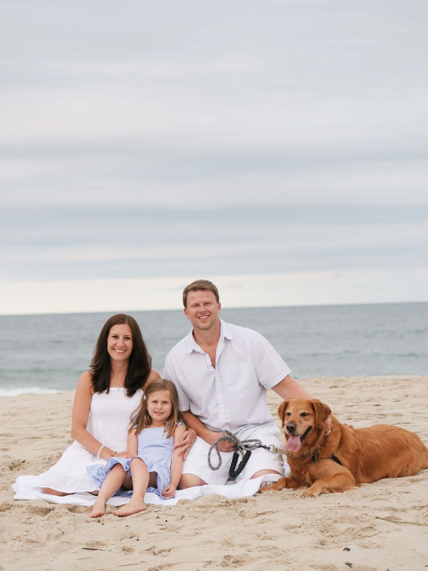 Family with young daughter and golden retriever posing on a sandy beach together.