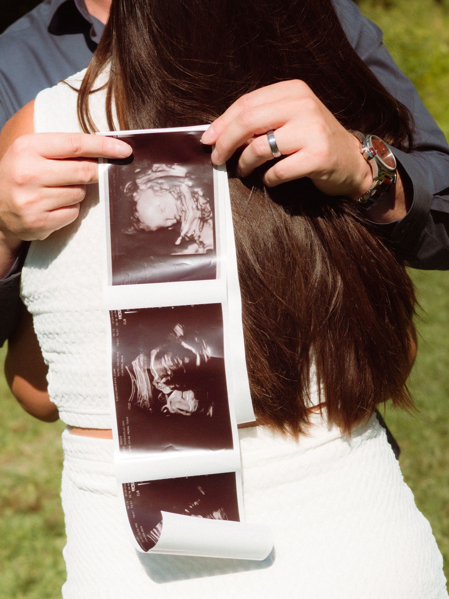 Couple holding ultrasound strip photos behind womans dark hair during maternity photo session.