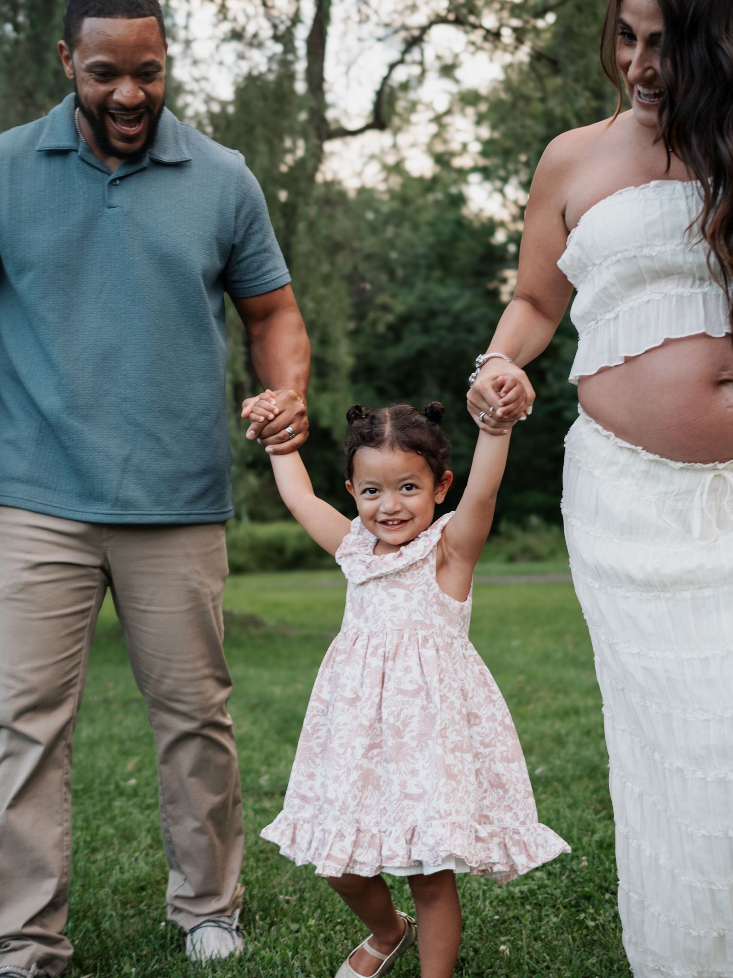 Happy toddler girl in floral dress holds parents hands while walking in green park.