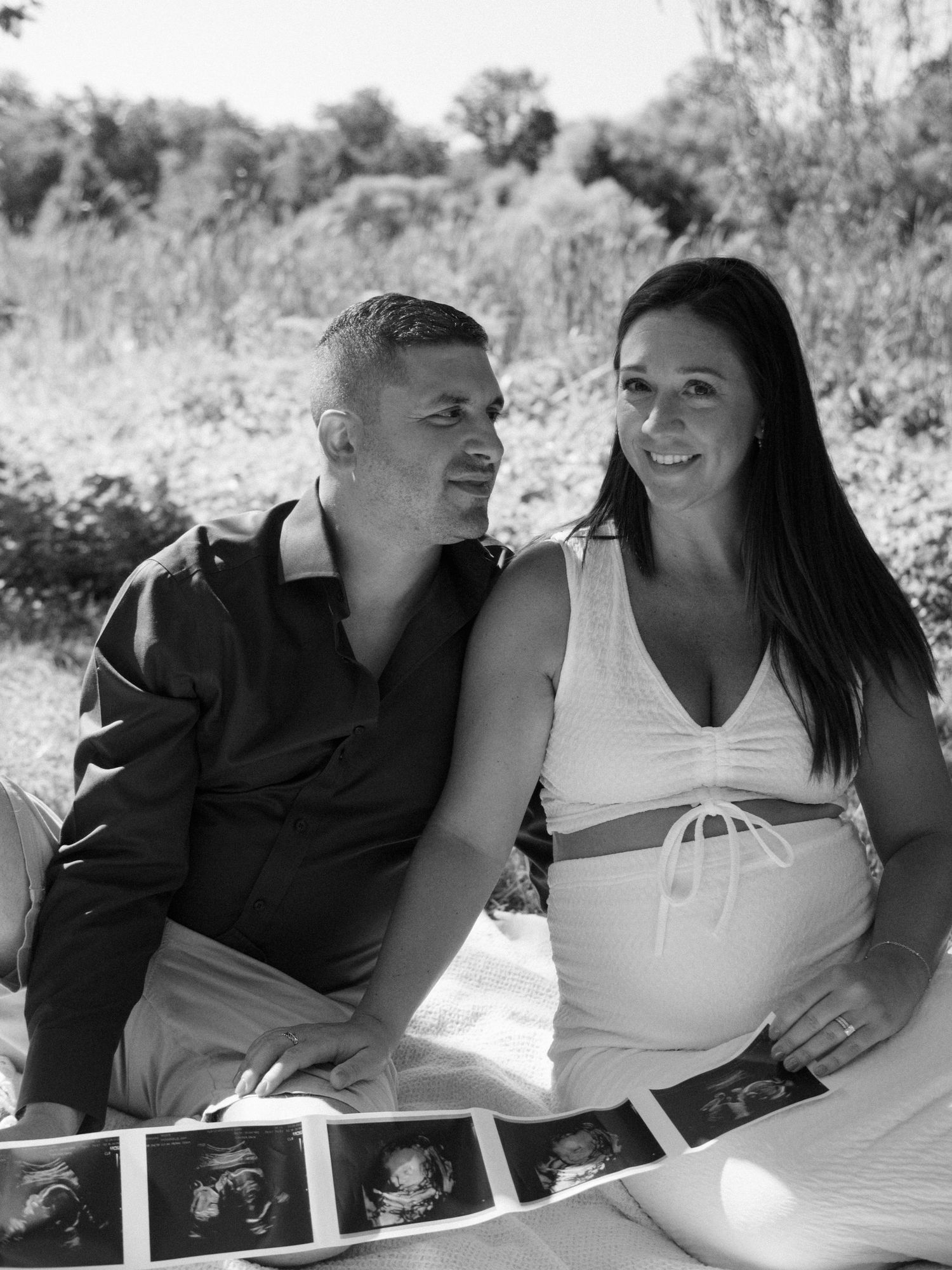 Black and white photo of expecting couple sitting outdoors holding ultrasound photos.
