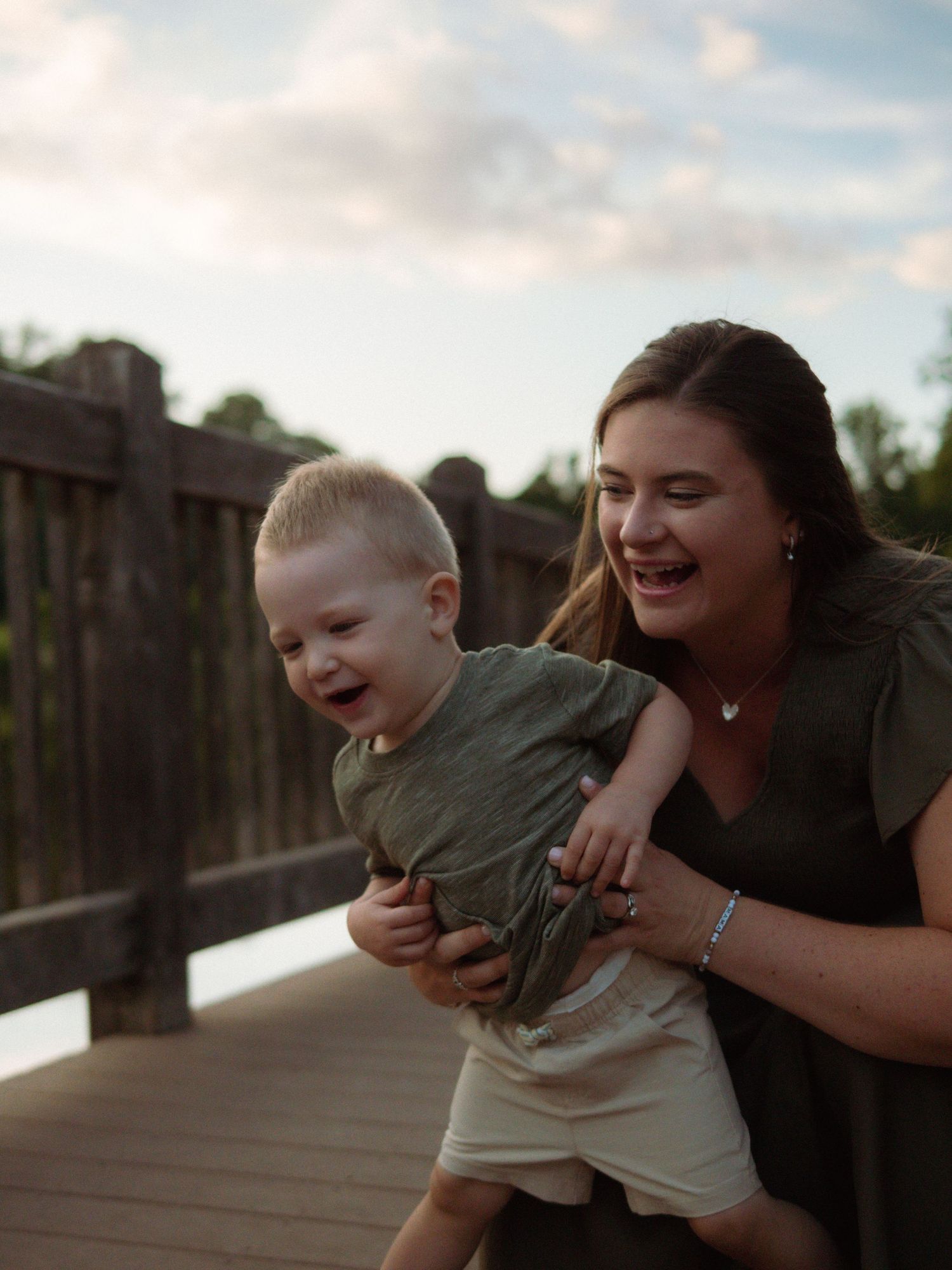 A laughing mother holds her giggling toddler son on a wooden bridge at sunset.