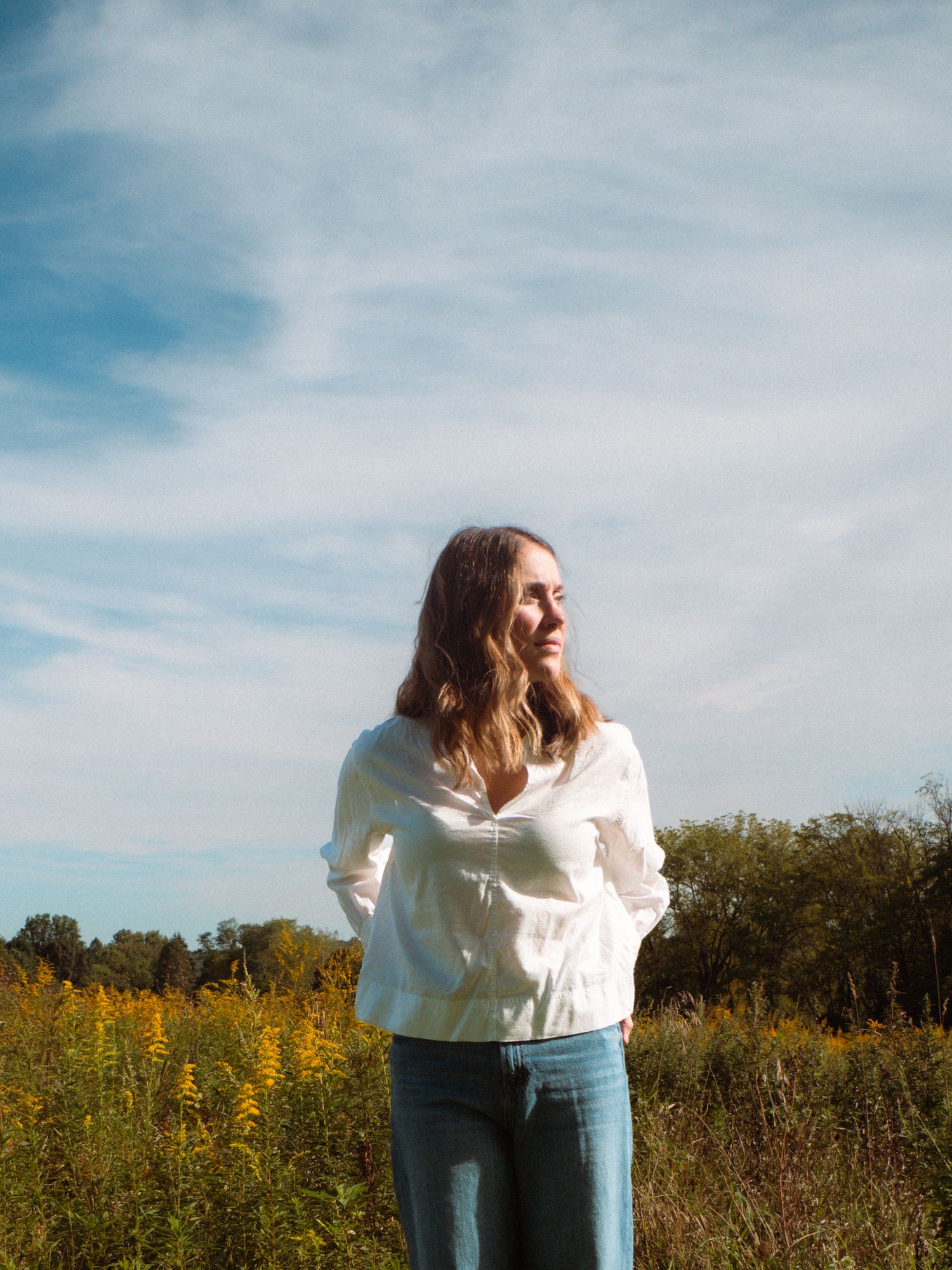 Young woman standing in golden wildflower field under blue sky wearing white blouse and jeans.