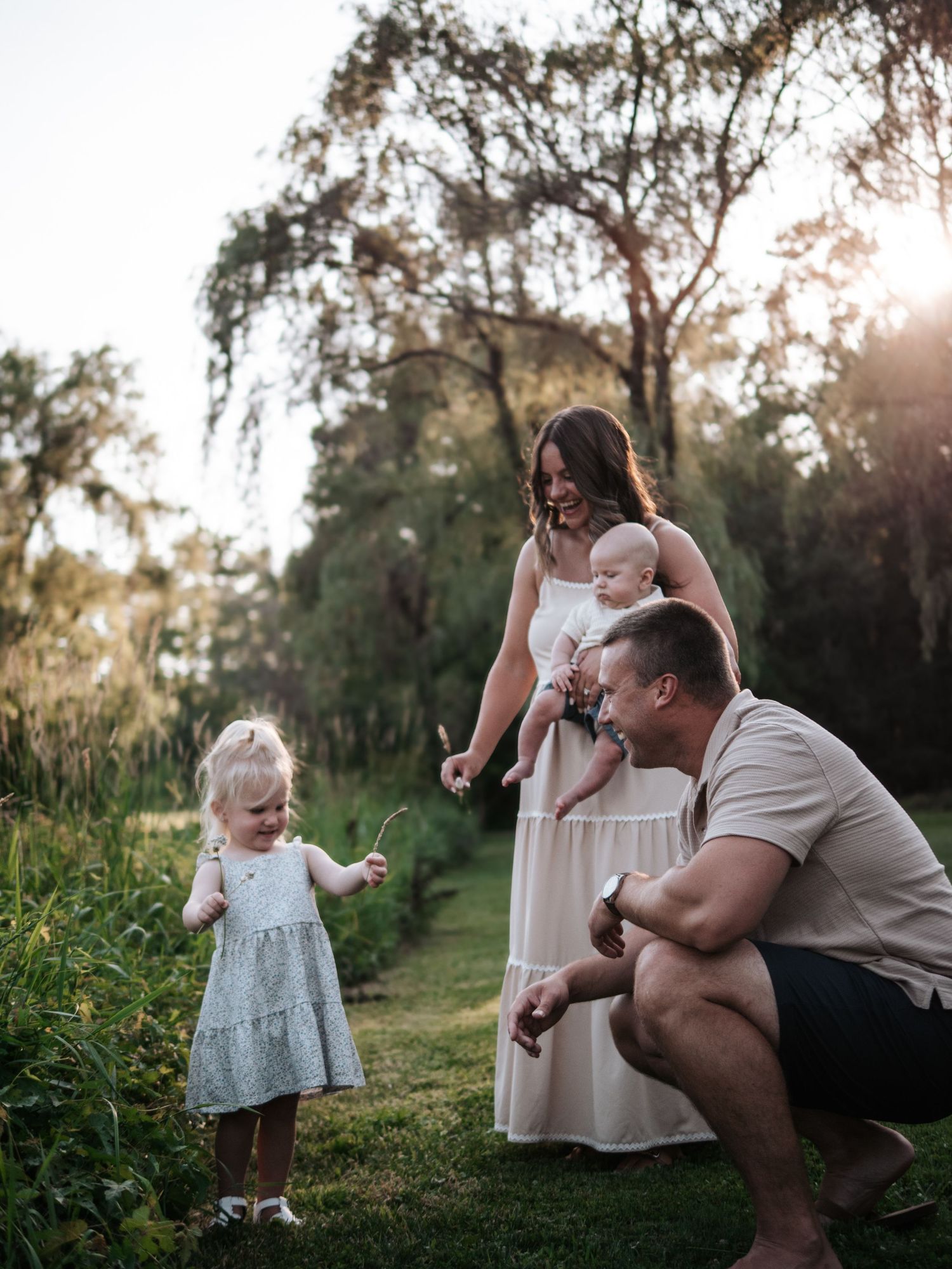 Father crouching to interact with toddler daughter while mother holds baby outdoors.