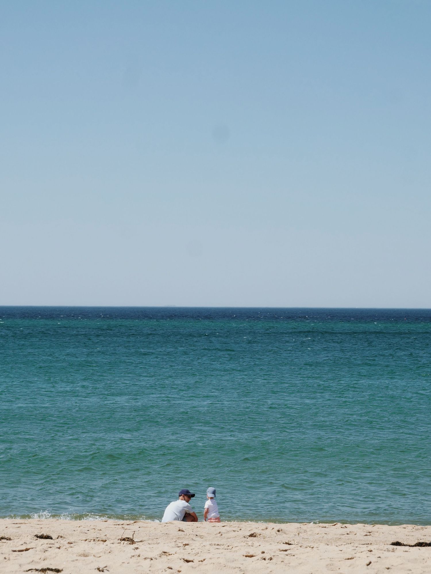 Parent and child sitting together on a sunny sandy beach by the ocean.