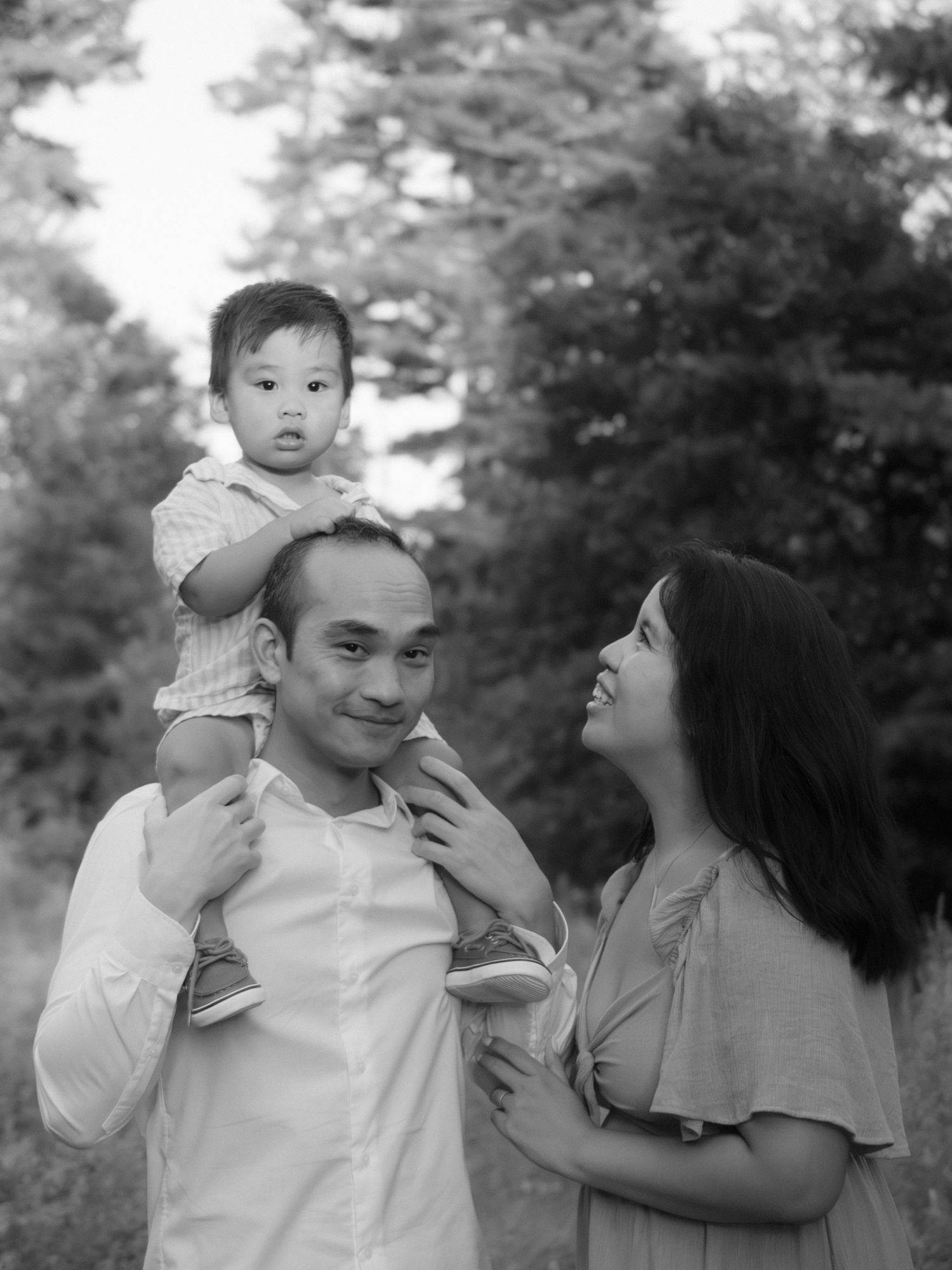 Black and white portrait of a father carrying his toddler on his shoulders with mother smiling.