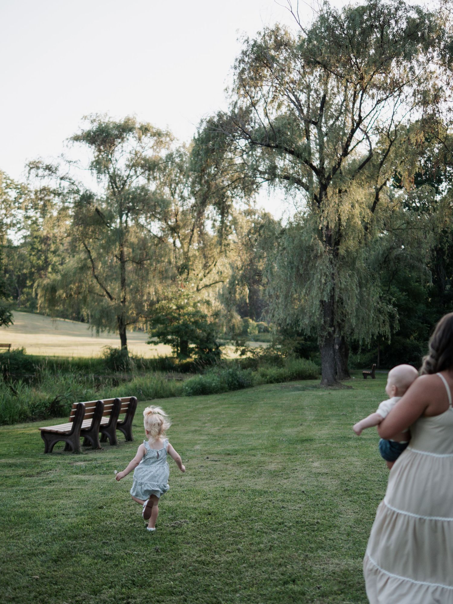 Young girl running toward mother holding baby in a scenic green park.