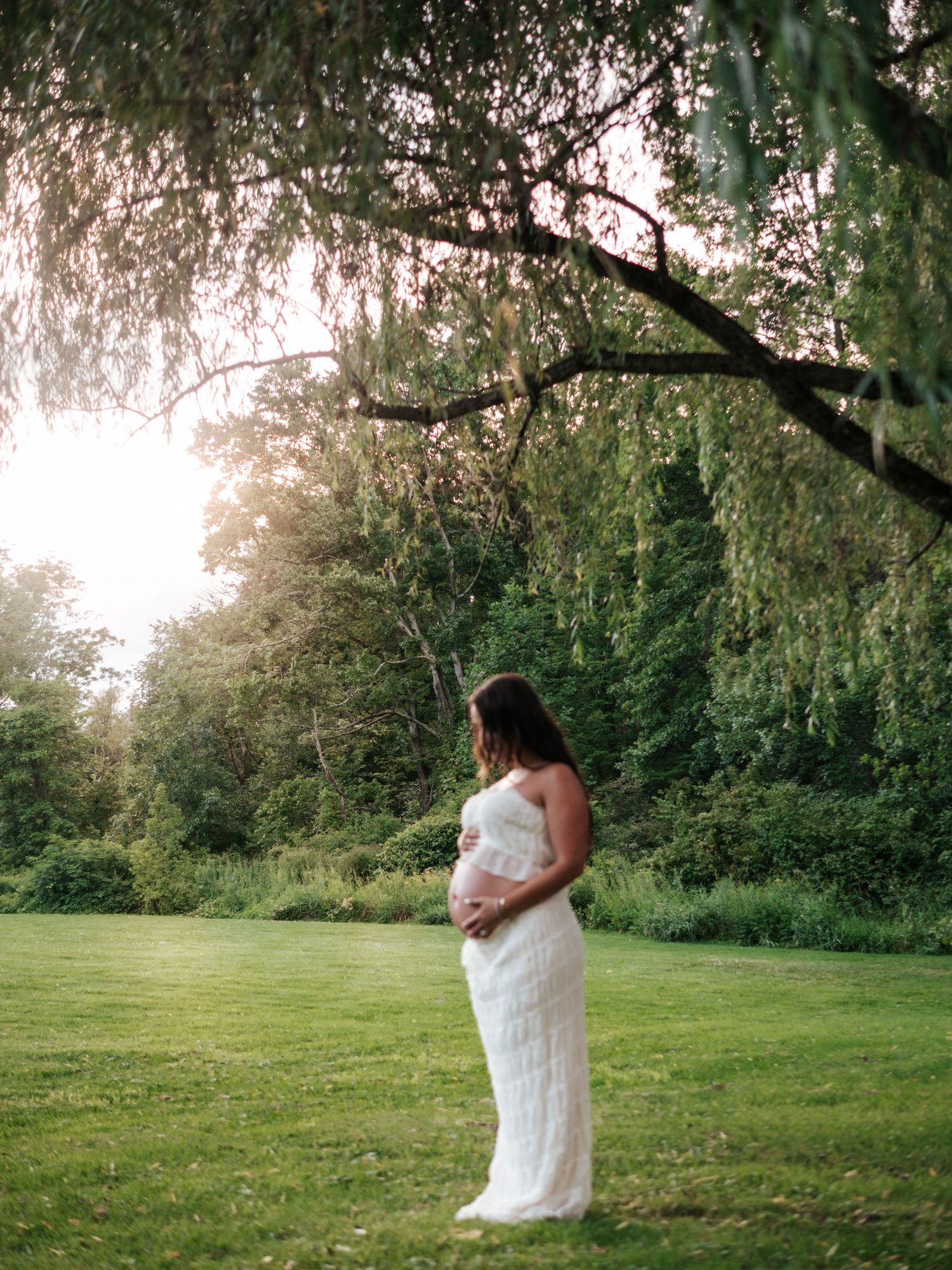 Pregnant woman in white dress standing under willow tree at golden hour in peaceful green park.