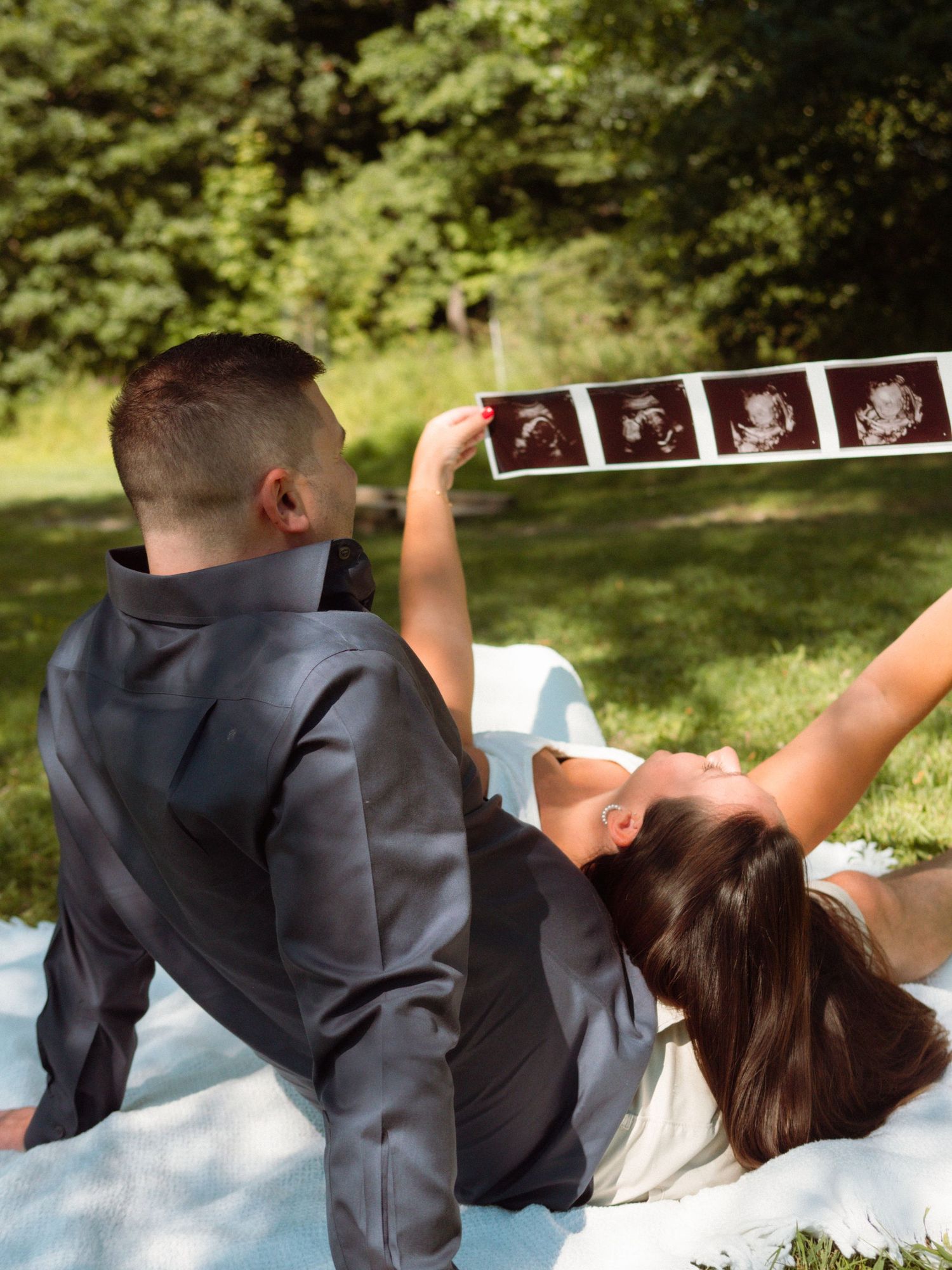 Expecting couple lying on grass holding ultrasound strip photos up in sunny park.