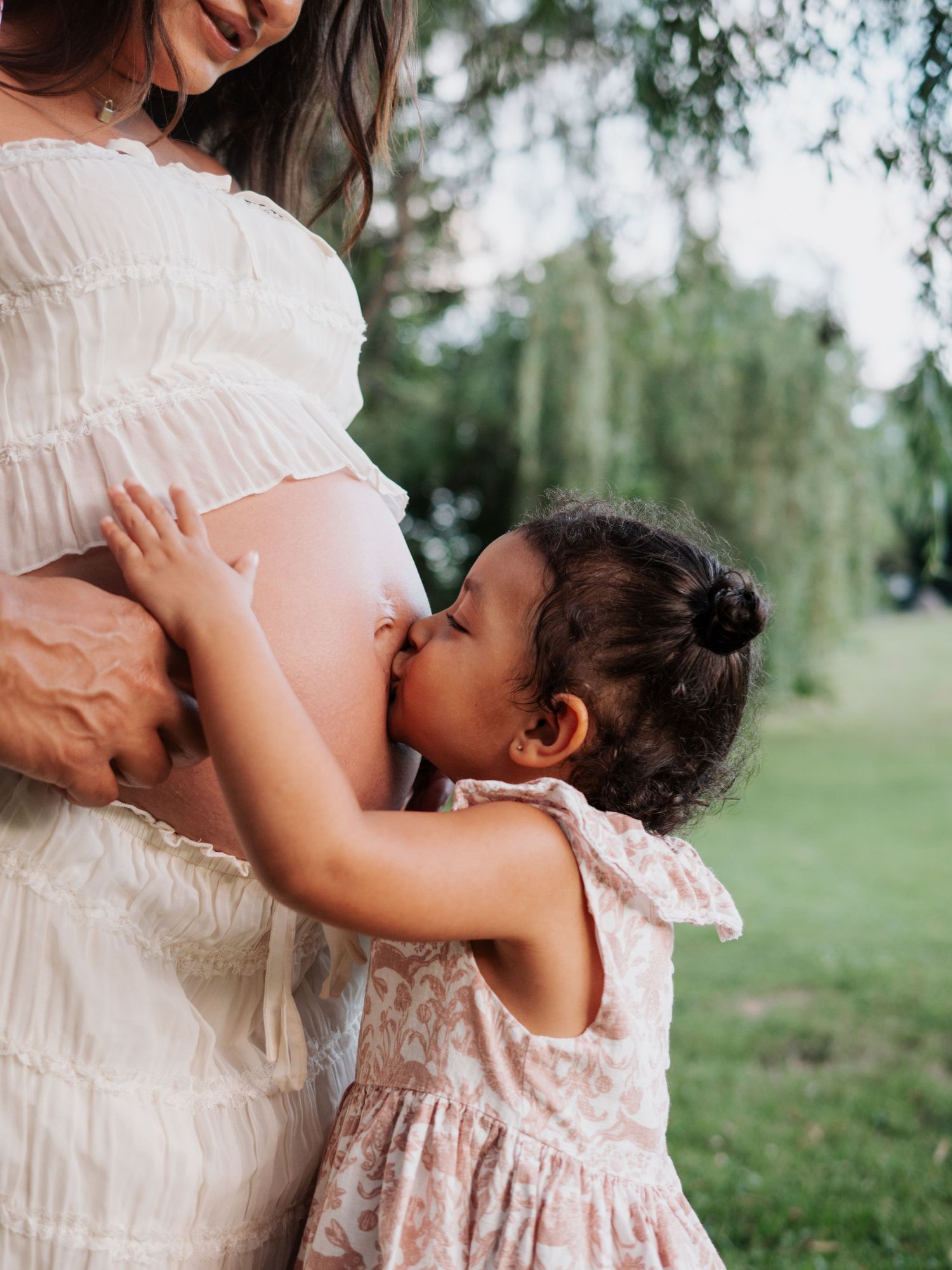 Toddler girl in floral dress kissing pregnant mothers bare belly outdoors near willow tree.