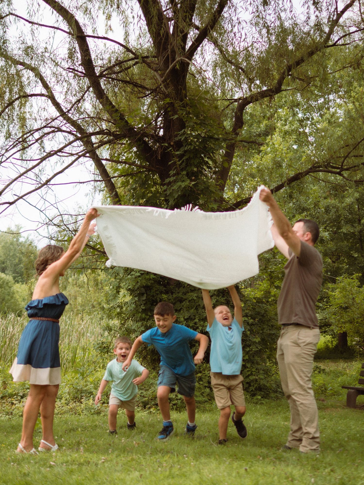 Family of five plays with a white blanket under a large tree in a lush park.