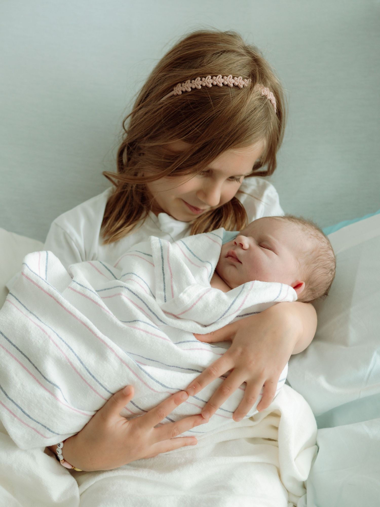 A young girl tenderly holds a swaddled newborn baby in a hospital setting.