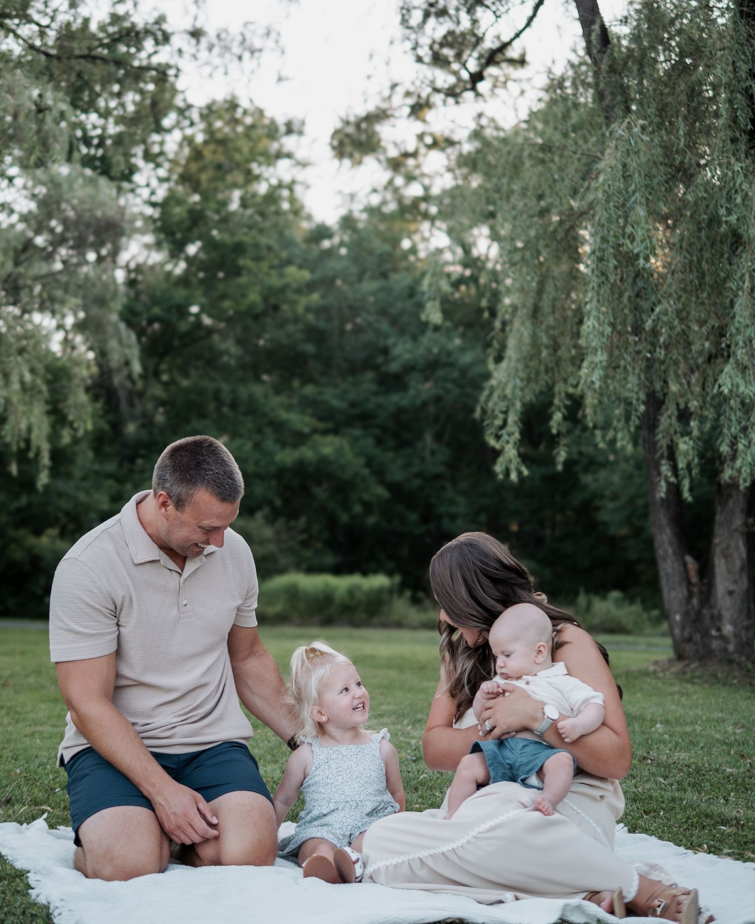 Happy family of four sitting on white blanket in park, parents smiling at toddler girl and baby in golden light.