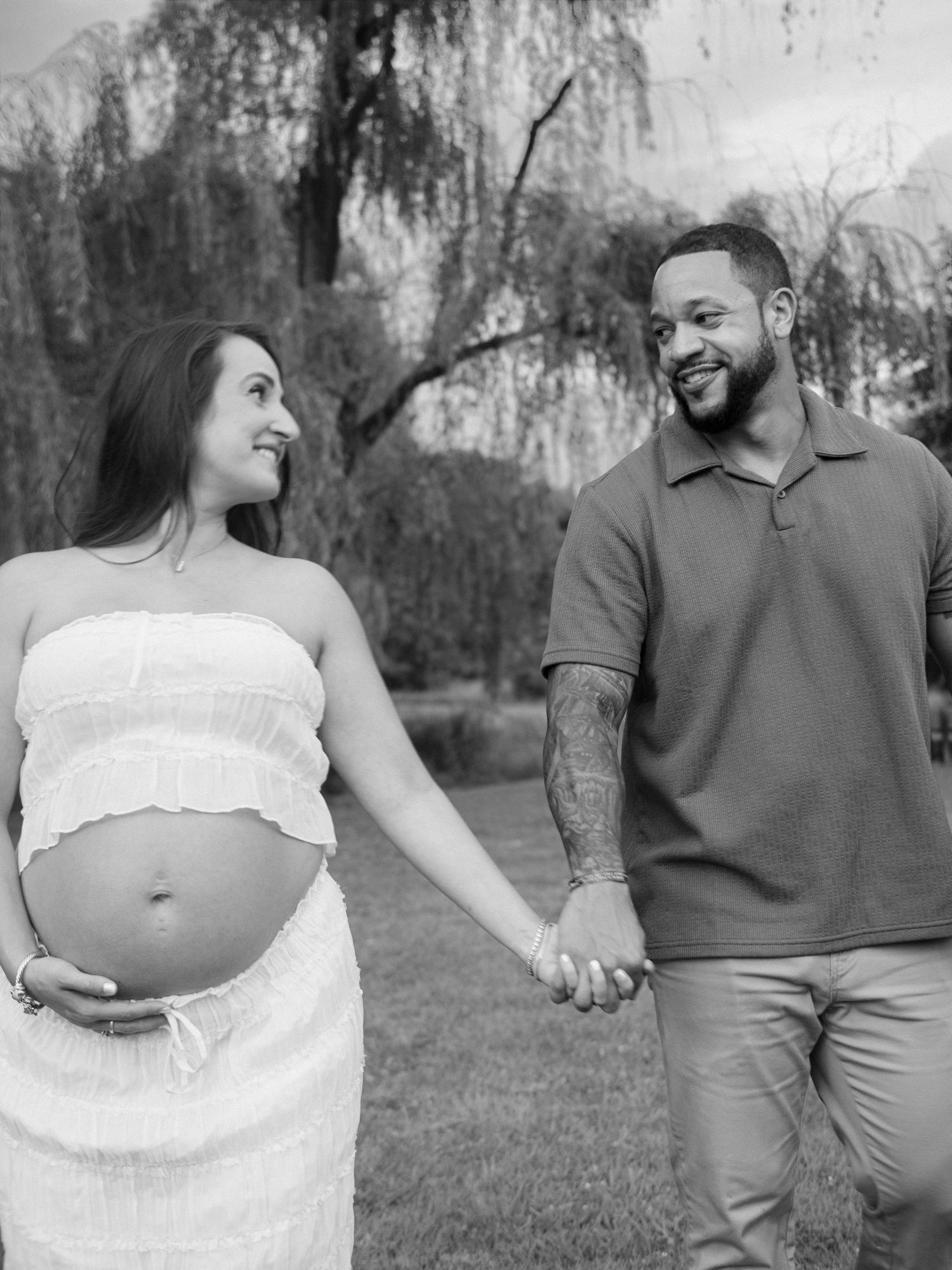 Black and white photo of smiling pregnant couple holding hands and gazing at each other outdoors.