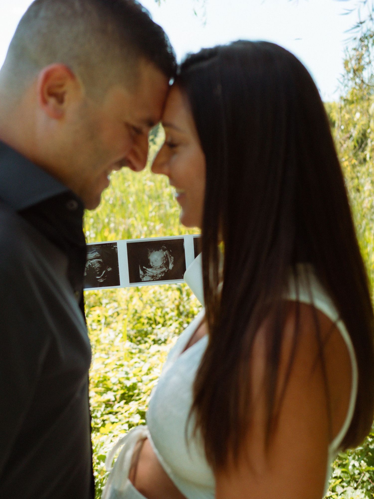 Expecting couple touching foreheads tenderly while holding ultrasound photos between them outdoors.