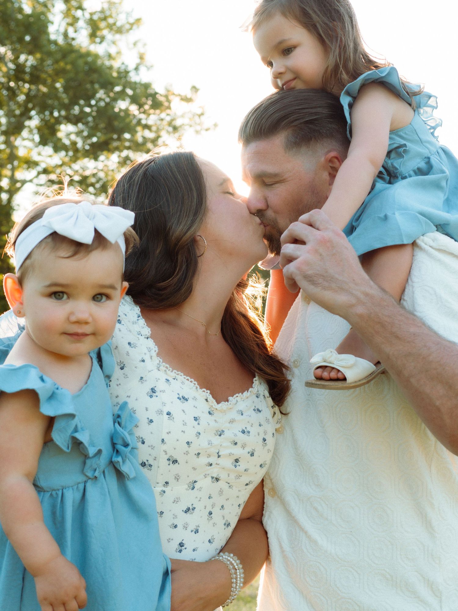 Family photo with parents kissing while holding two young daughters in matching blue dresses outdoors.