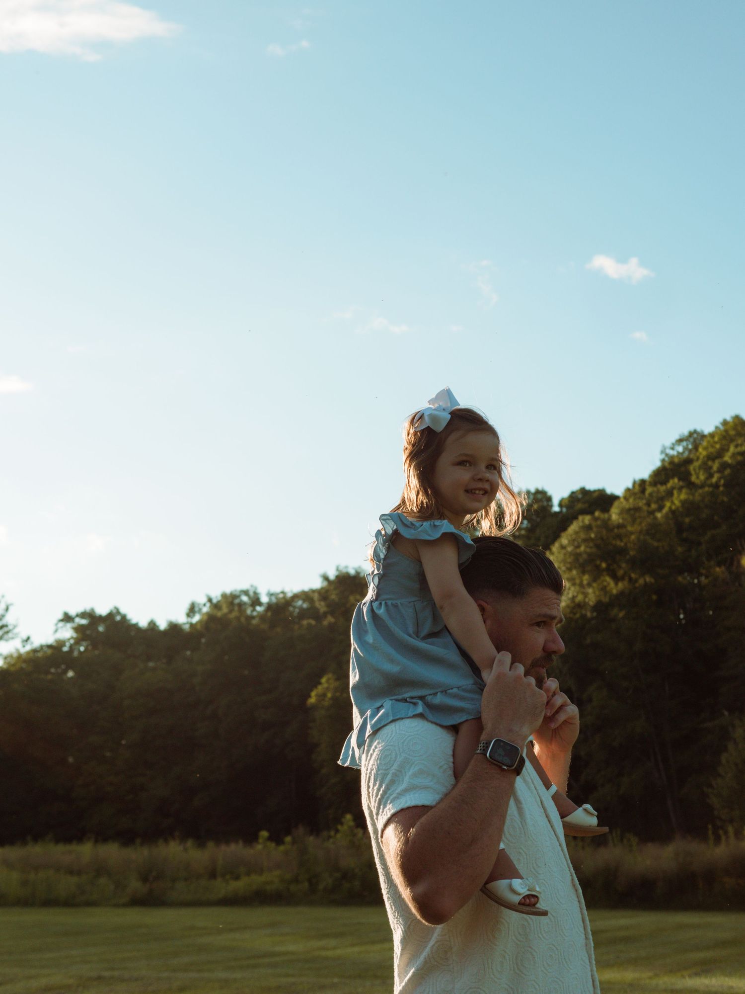 Little girl in blue dress sitting on father's shoulders in a sunny park.