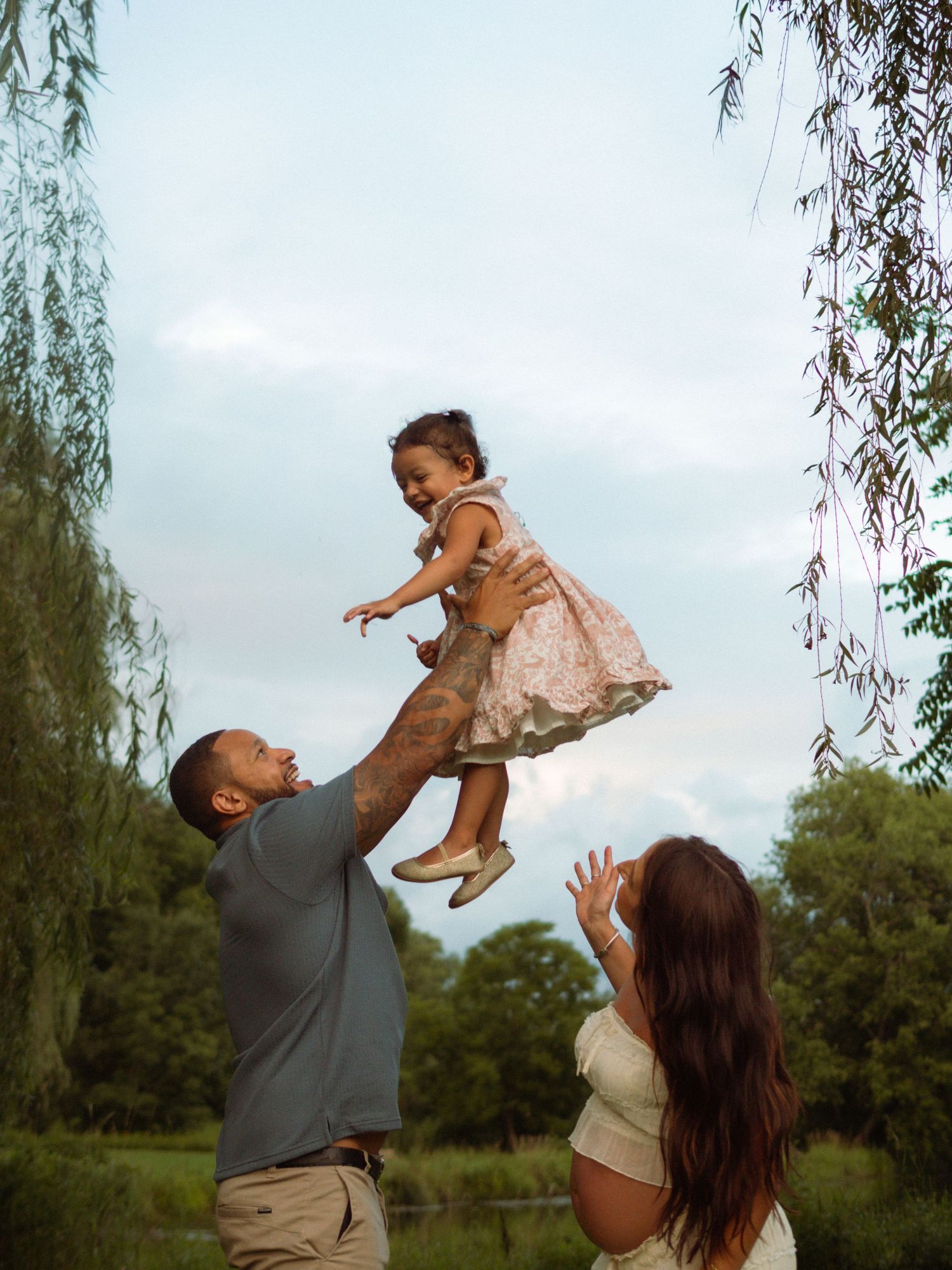 Father tossing laughing toddler girl in floral dress into the air outdoors.