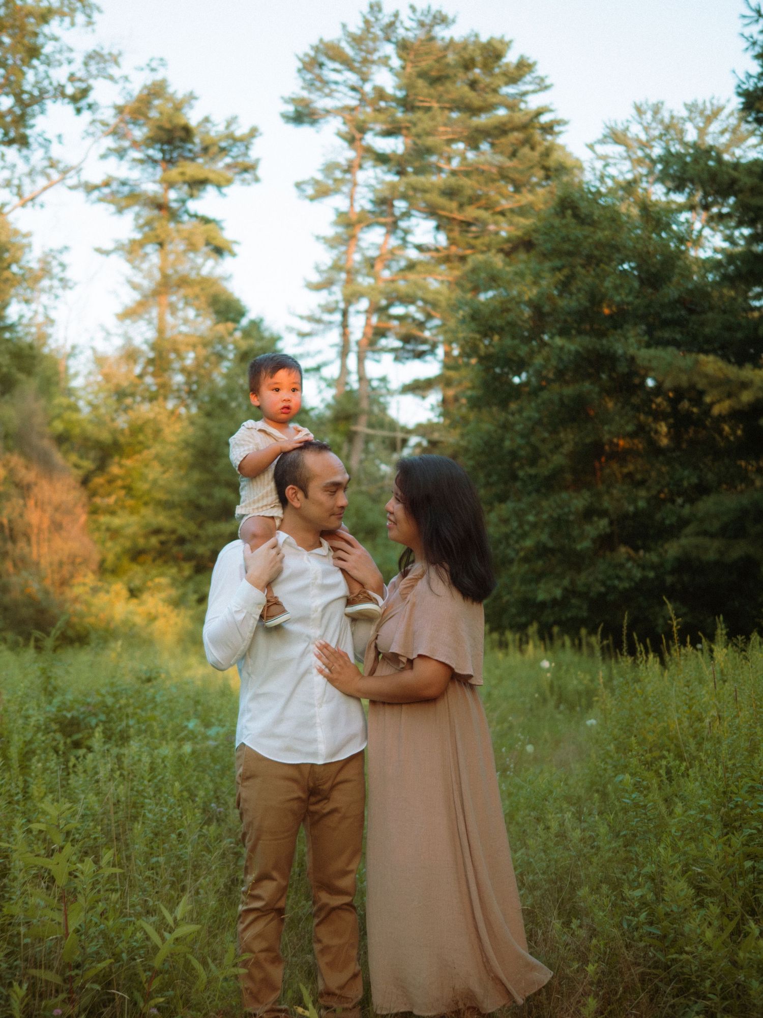 A family of three poses in a lush green meadow with tall pine trees.